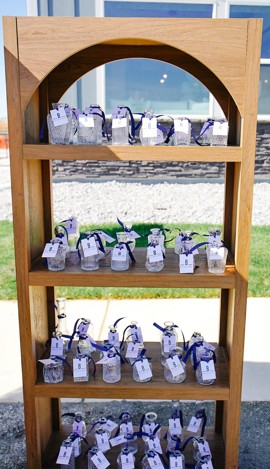 Bespoke wooden arch shelf displaying vintage-style glass bud vases tied with purple ribbons and custom event tags. Designed as a take-home floral favor station for a mobile Bloom Bar event in Denver and Highlands Ranch, Colorado.