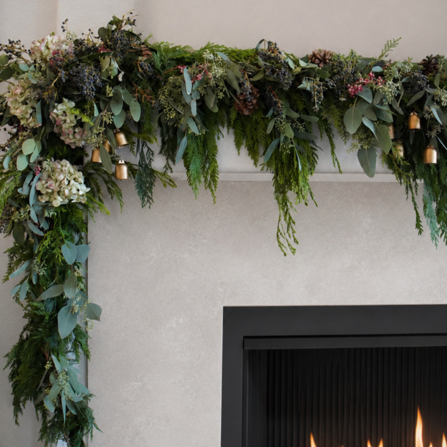 Green holiday garland with sprinkling of pinecones, berries, and small gold bells hangs over a fireplace.