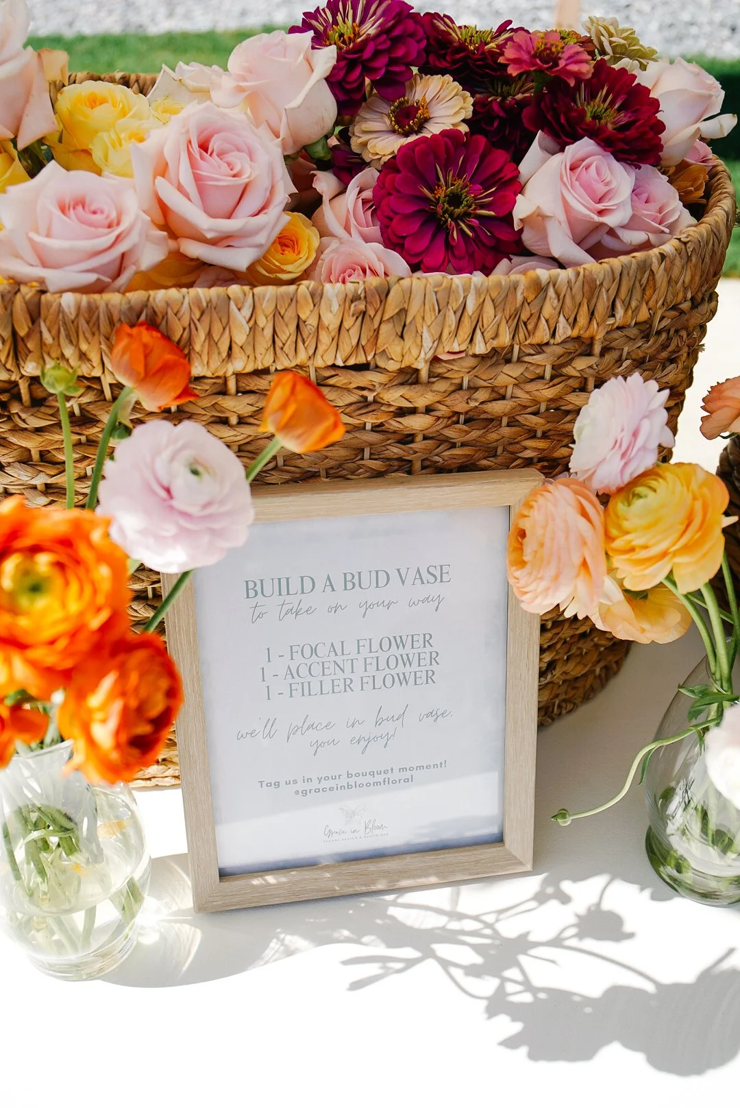 Interactive 'Build a Bud Vase' instructional sign framed beside vibrant orange ranunculus, blush roses, and deep magenta zinnias in woven baskets. A perfect DIY flower bar activity and favor station for bridal showers, baby showers, and private event