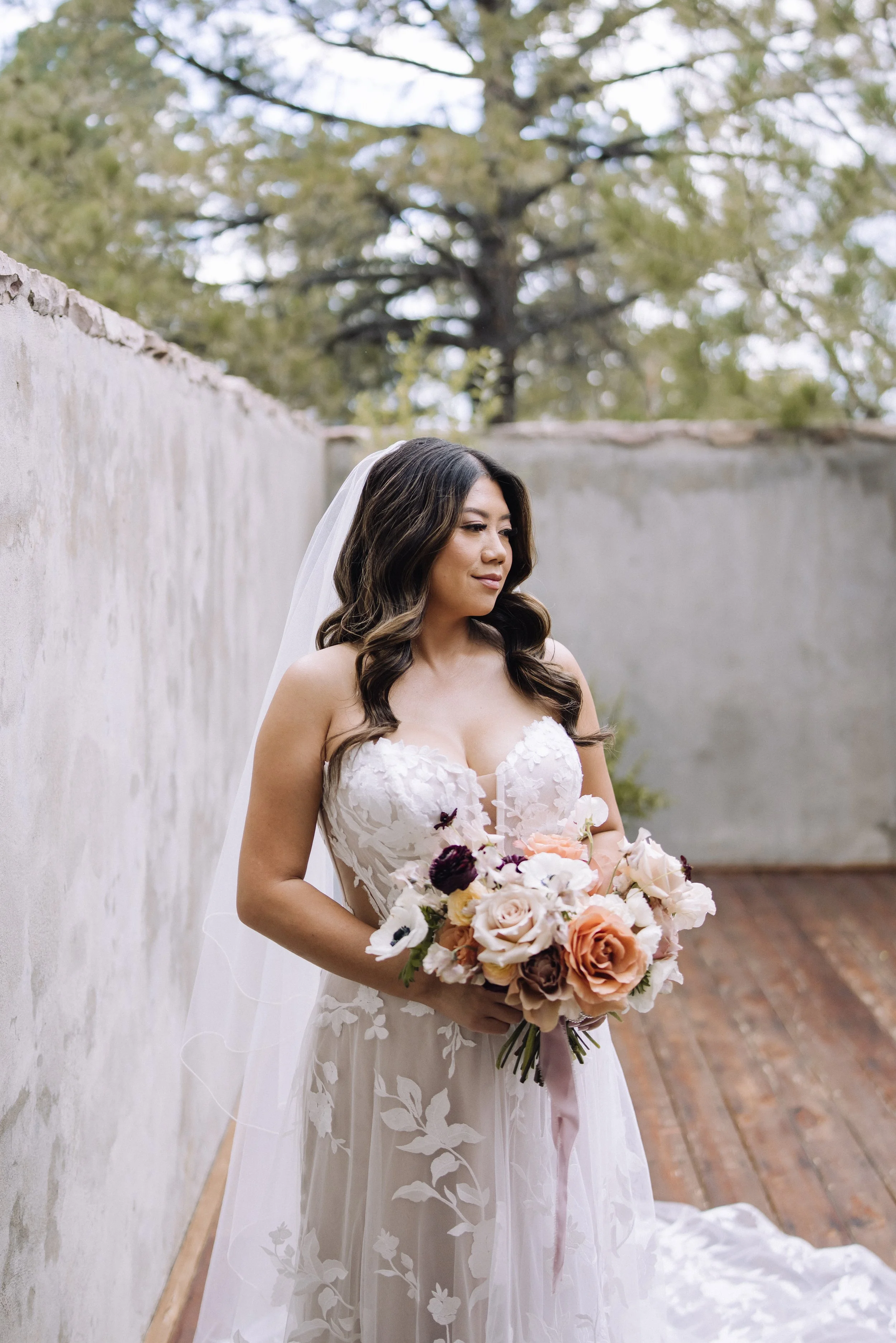 Bride holding a blush, peach, and deep plum bridal bouquet while posing outdoors at The Oaks wedding venue in Castle Rock, CO, designed by Grace in Bloom Floral.