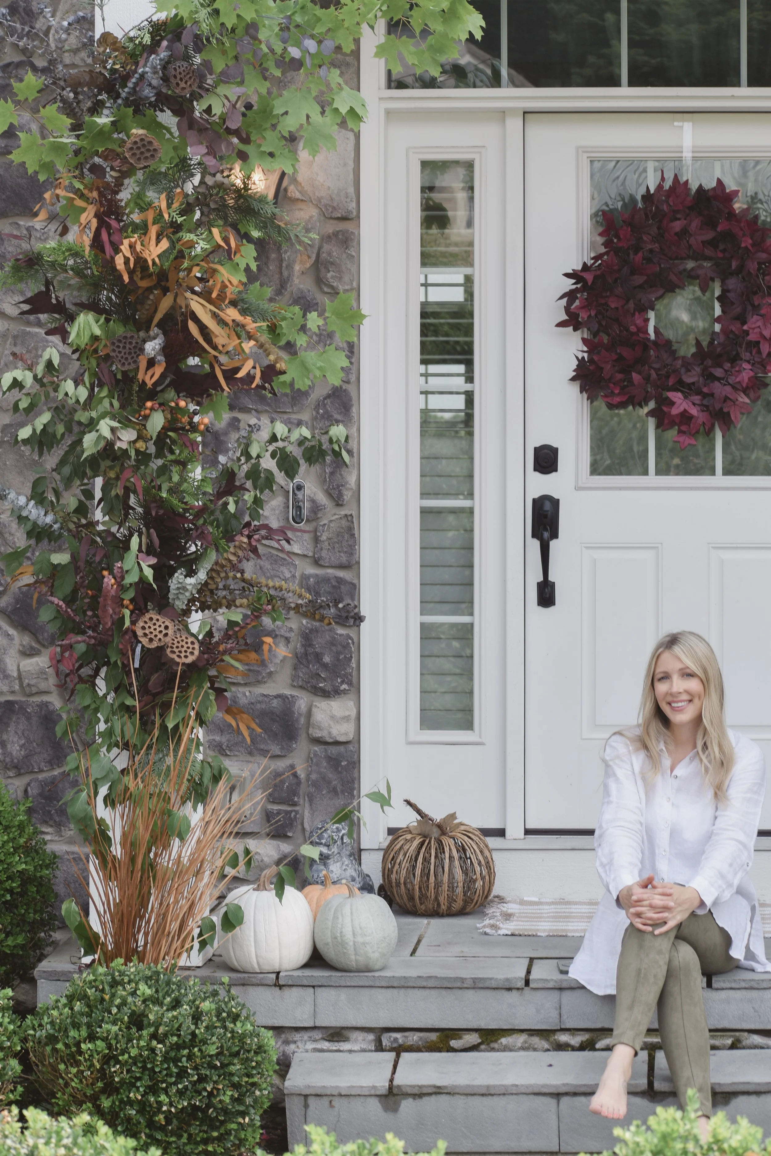 A woman sitting on concrete steps outside a house decorated for fall with pumpkins, gourds, and seasonal plants, smiling at the camera.