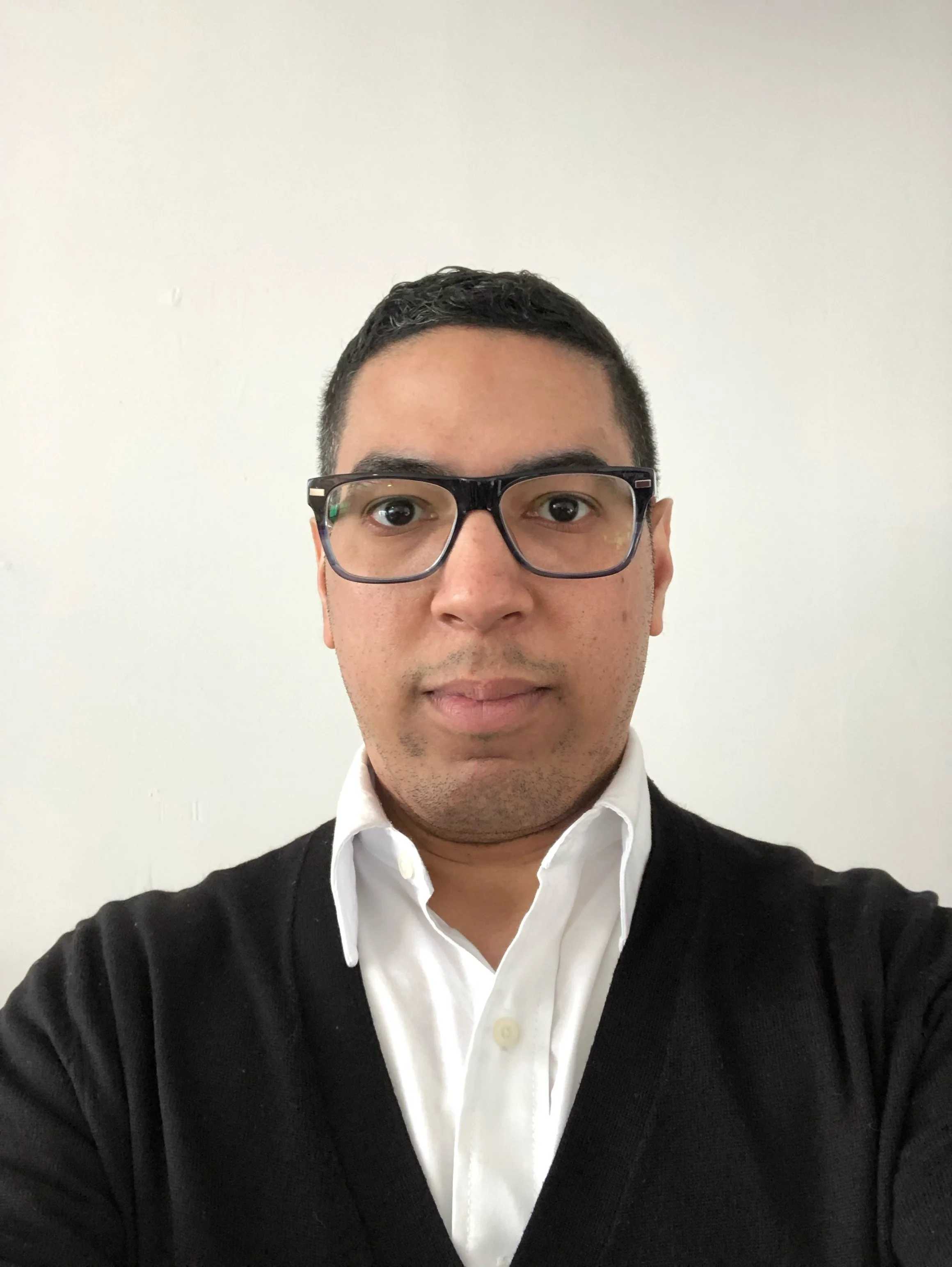 A man wearing glasses, a white shirt, and a black sweater, taking a selfie against a plain white wall.