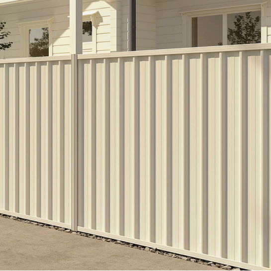Close-up of a beige metal privacy fence in front of a white house with windows.