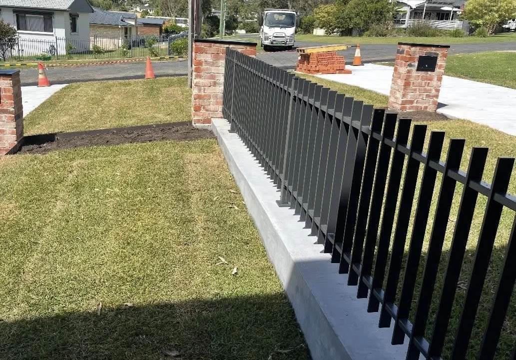 A black metal fence running along a concrete sidewalk, with brick columns on each side, surrounding a lawn and a garden bed, in a residential area with houses and trees in the background.
