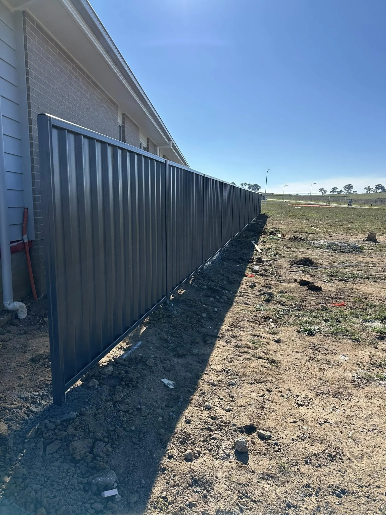 Metal fence installed along the side of a house on a dirt lot with some patches of grass, under a clear blue sky.