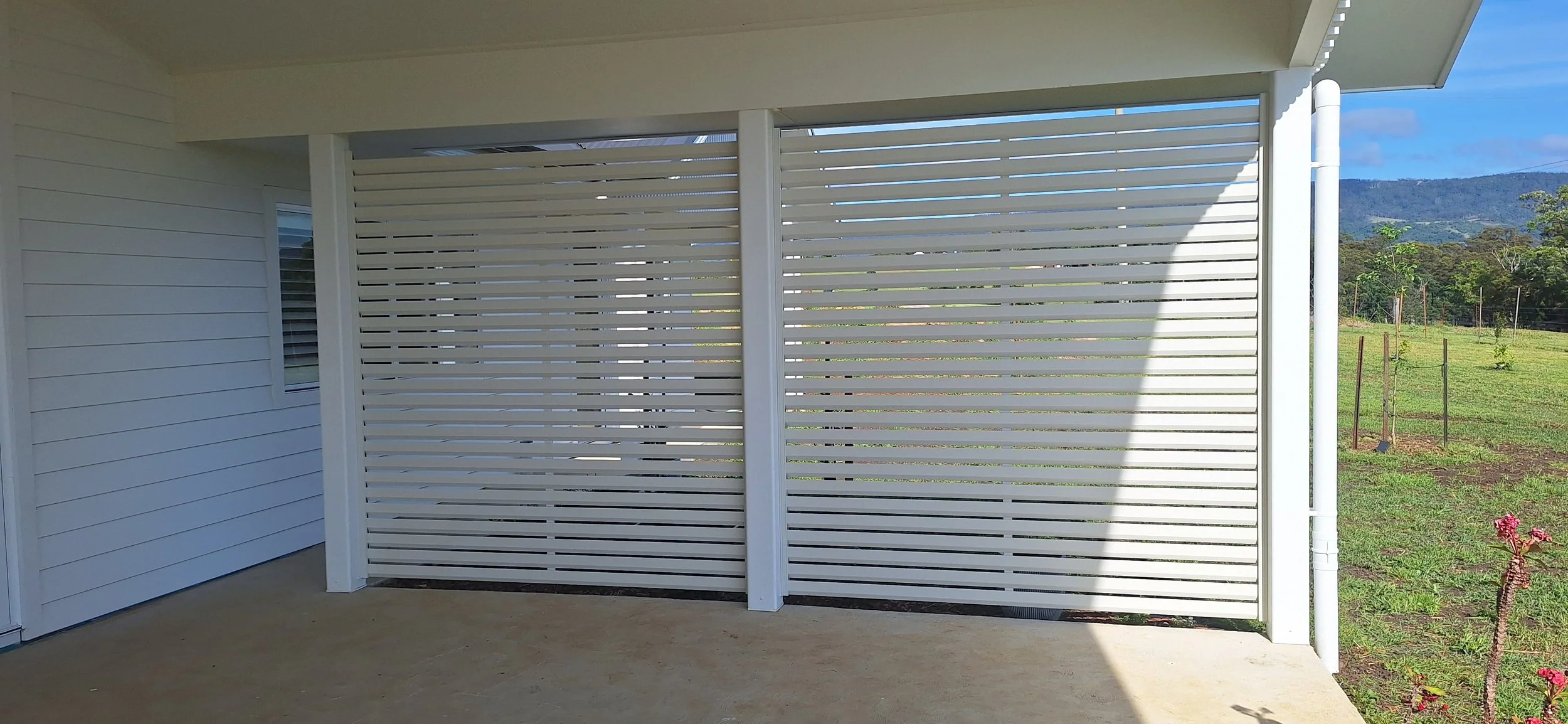 White horizontal slat privacy screen on an outdoor patio, with a view of a grassy field and trees in the background.