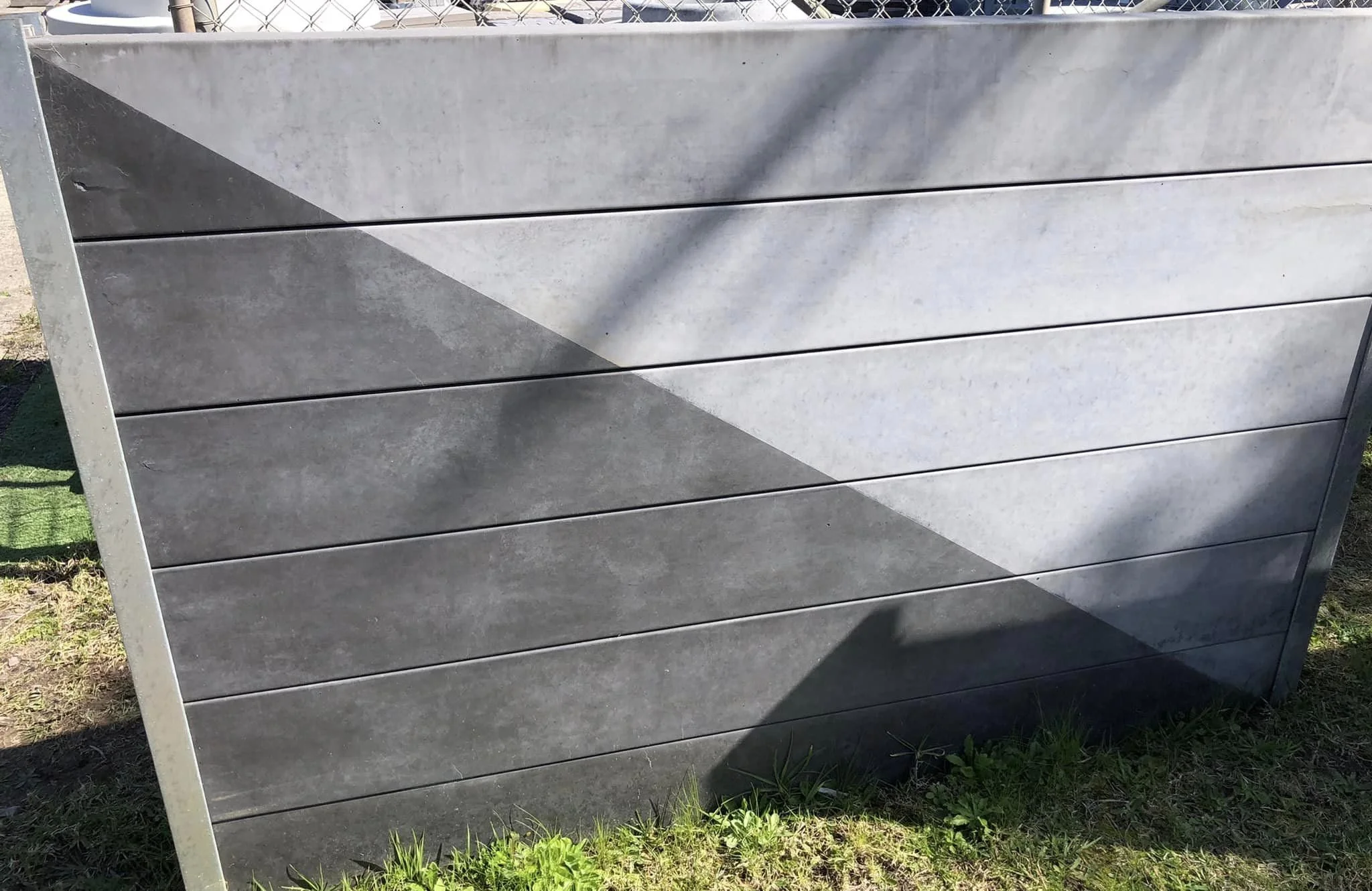 Close-up of a modern, rectangular concrete planter box with horizontal slats, situated outdoors on grass with shadow cast on it.