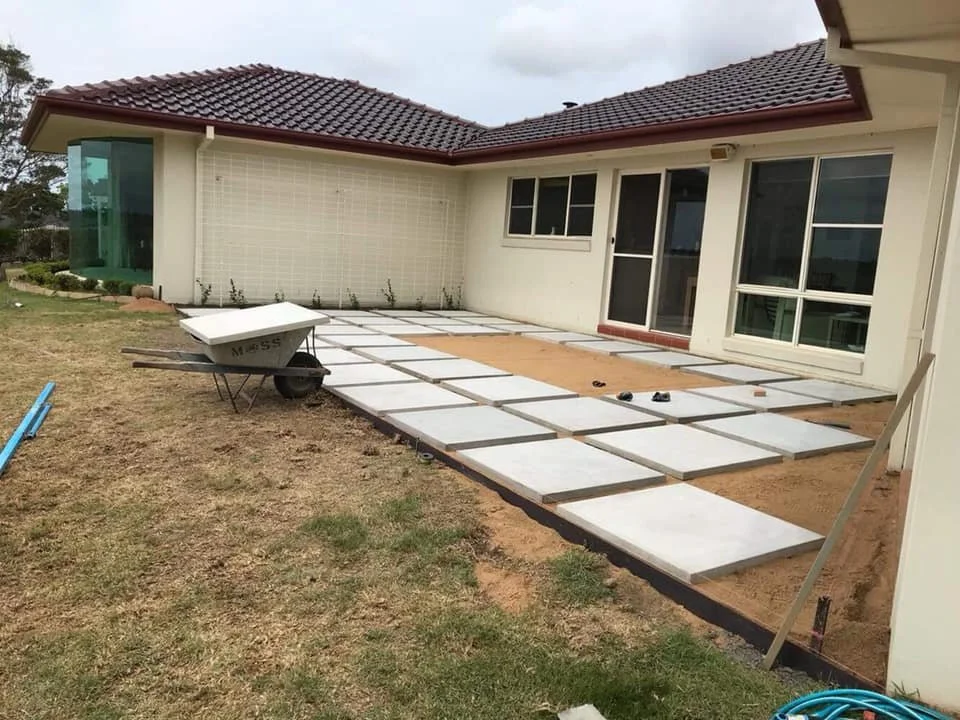 Home exterior undergoing patio construction with concrete slabs laid out on a dirt surface, a wheelbarrow, and some tools.