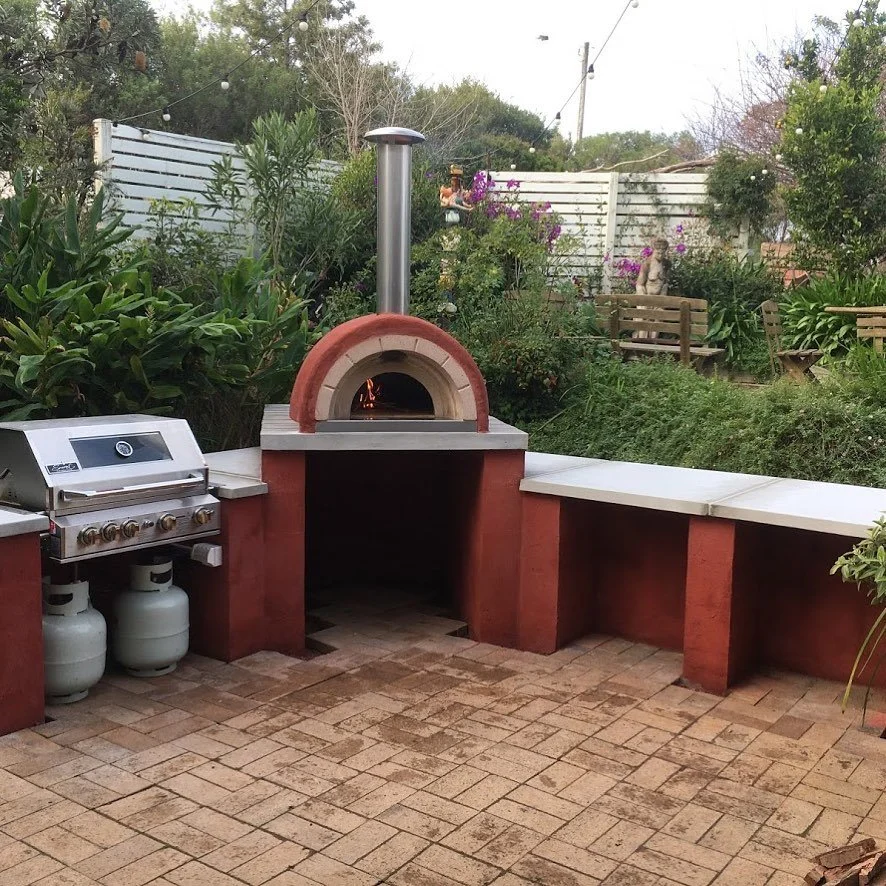 Outdoor backyard with brick barbecue oven, side benches, gas grill with propane tanks, surrounded by greenery and garden furniture.