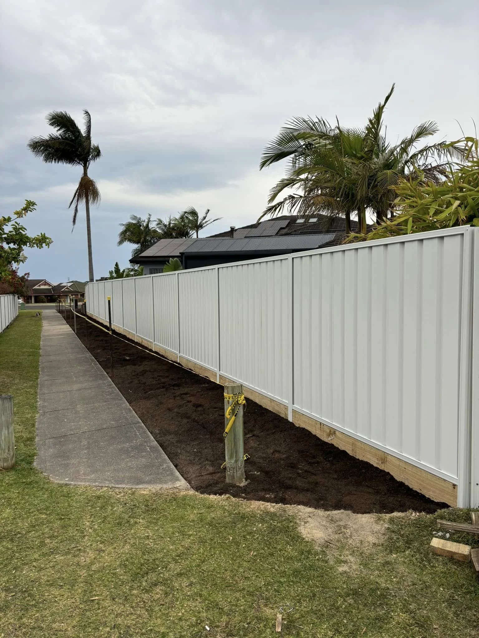 Sidewalk next to a white fence with a sidewalk and grass in the foreground and a partially planted area with dark soil along the base of the fence. Palm trees and suburban houses are visible in the background under cloudy skies.