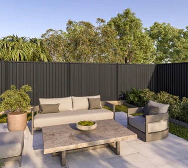 Outdoor patio with beige sofa, armchairs, coffee table, potted plant, and greenery, enclosed by a black fence, under a clear blue sky. Dark grey metal fence.