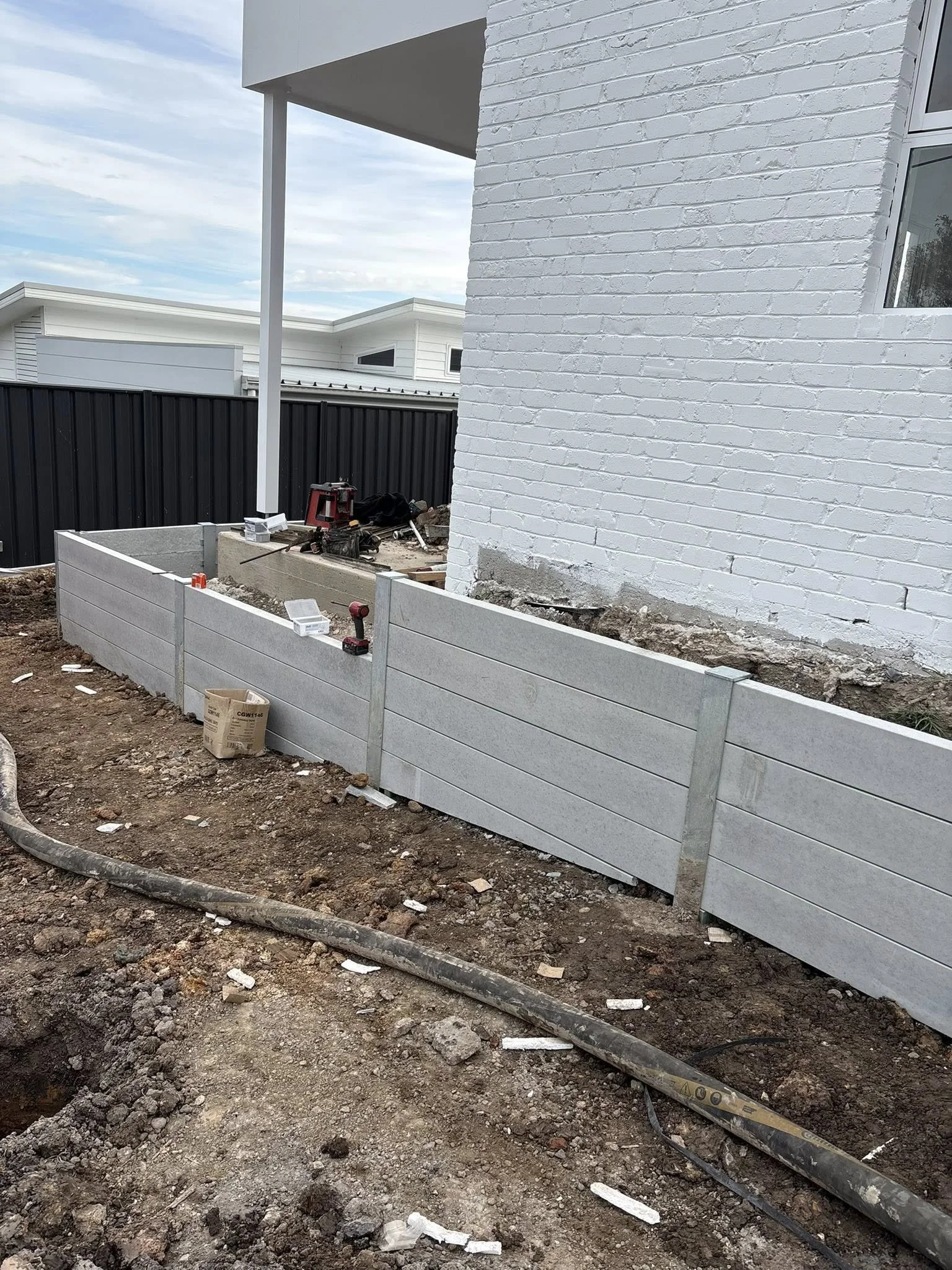 Construction site with a freshly installed gray fence along the side of a white brick house, with construction tools and materials on the ground and a partially dug trench in the dirt yard.