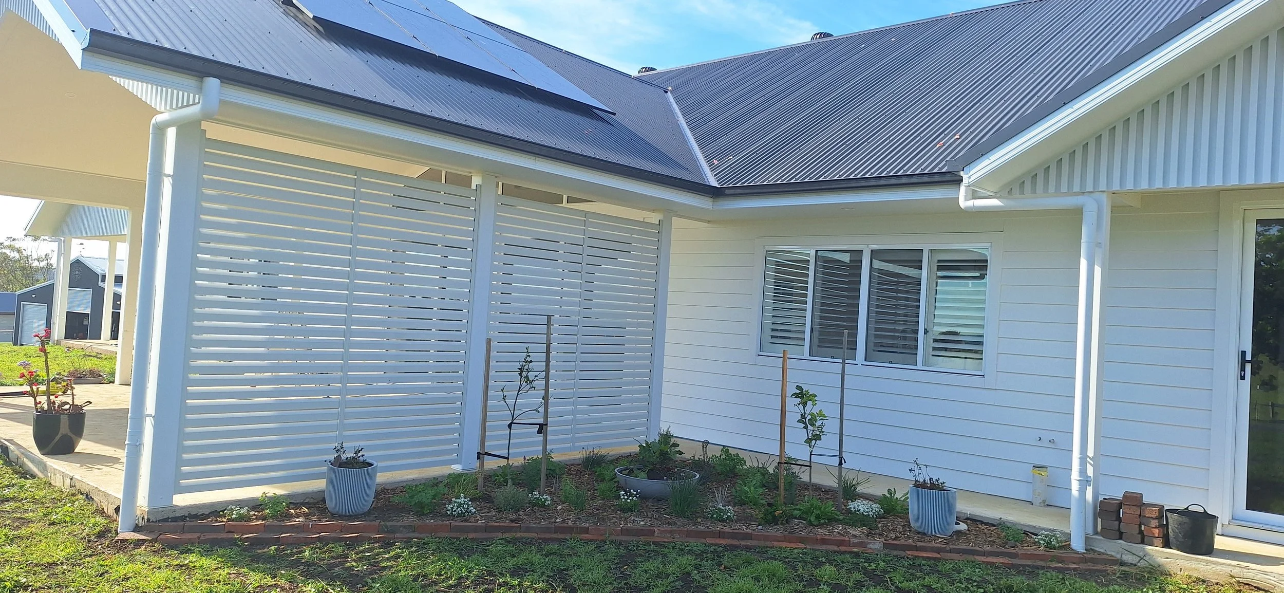 The image shows the exterior of a modern house with white siding and a black metal roof. There is a small garden with young plants and flowers in front of the house, along with potted plants on the ground. The house has large windows with white blinds and a sliding glass door. A white slatted privacy fence is attached to the house, and a few bricks and a black bucket are on the ground near the door.