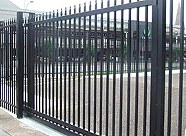 Black metal fence with vertical bars, situated outdoors on paved ground, with a baseball field in the background.