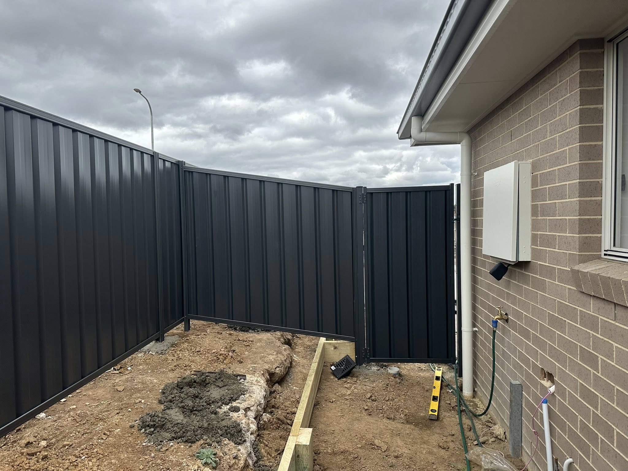 Backyard with a metal privacy fence and a brick house with outdoor piping and electrical boxes under a cloudy sky.