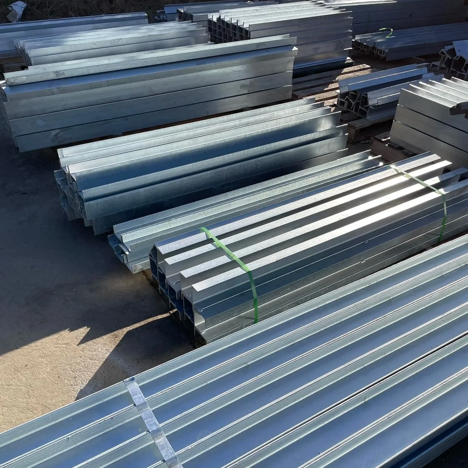 Stacks of metal construction beams and channels arranged outdoors on concrete ground.