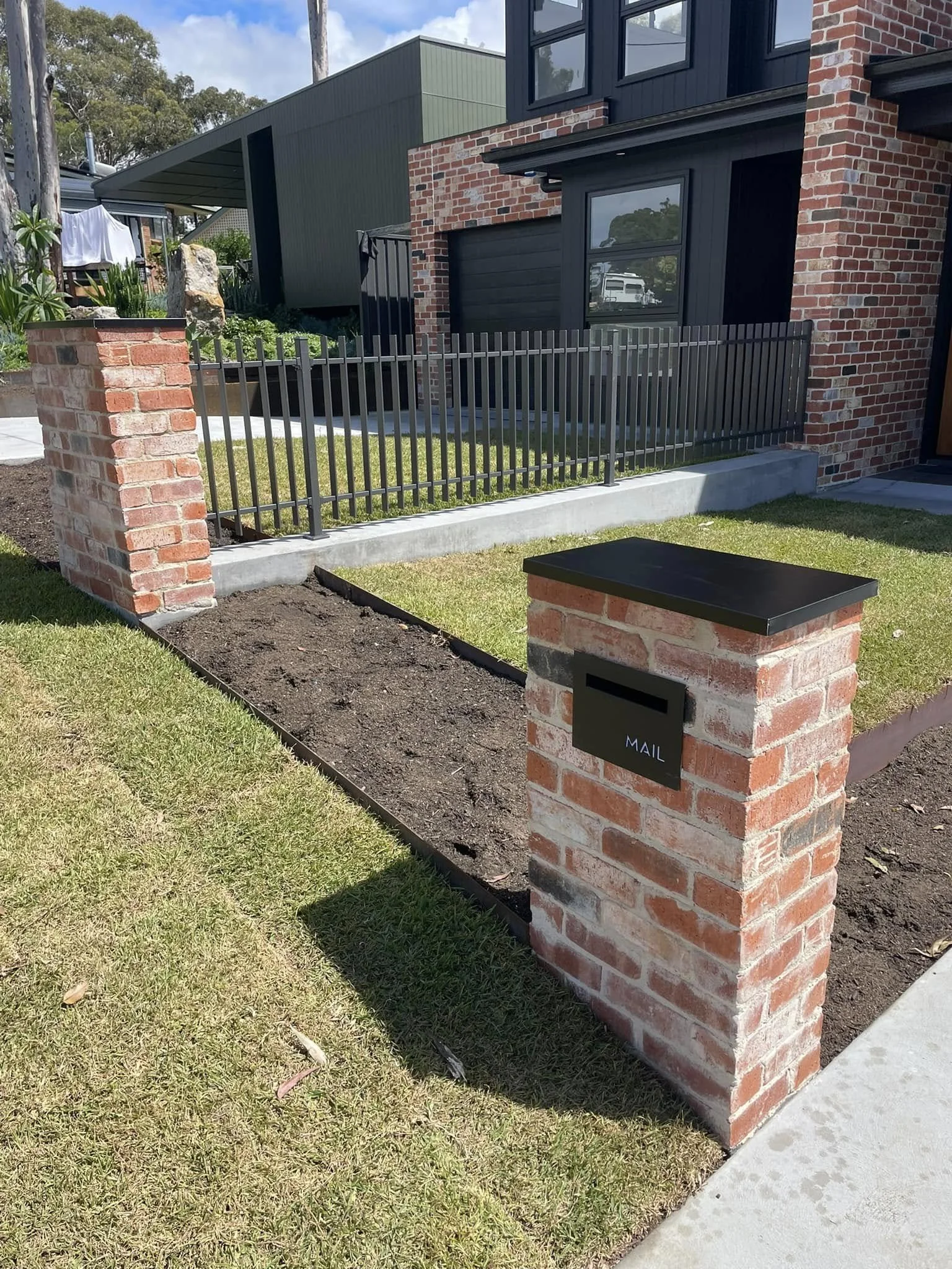 Front yard with brick mailbox labeled 'MAIL', garden bed with soil, and modern house with brick and dark-colored siding.