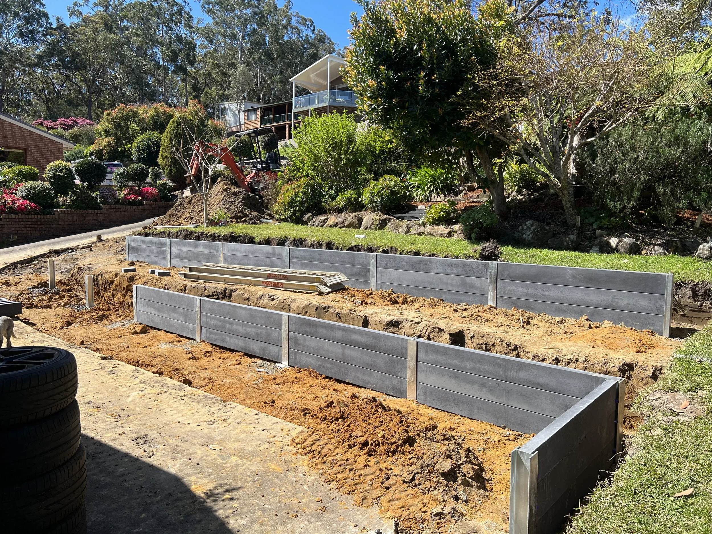 Construction site in a backyard with a trench, metal form panels, orange dirt, and construction tools, with trees and houses in the background.