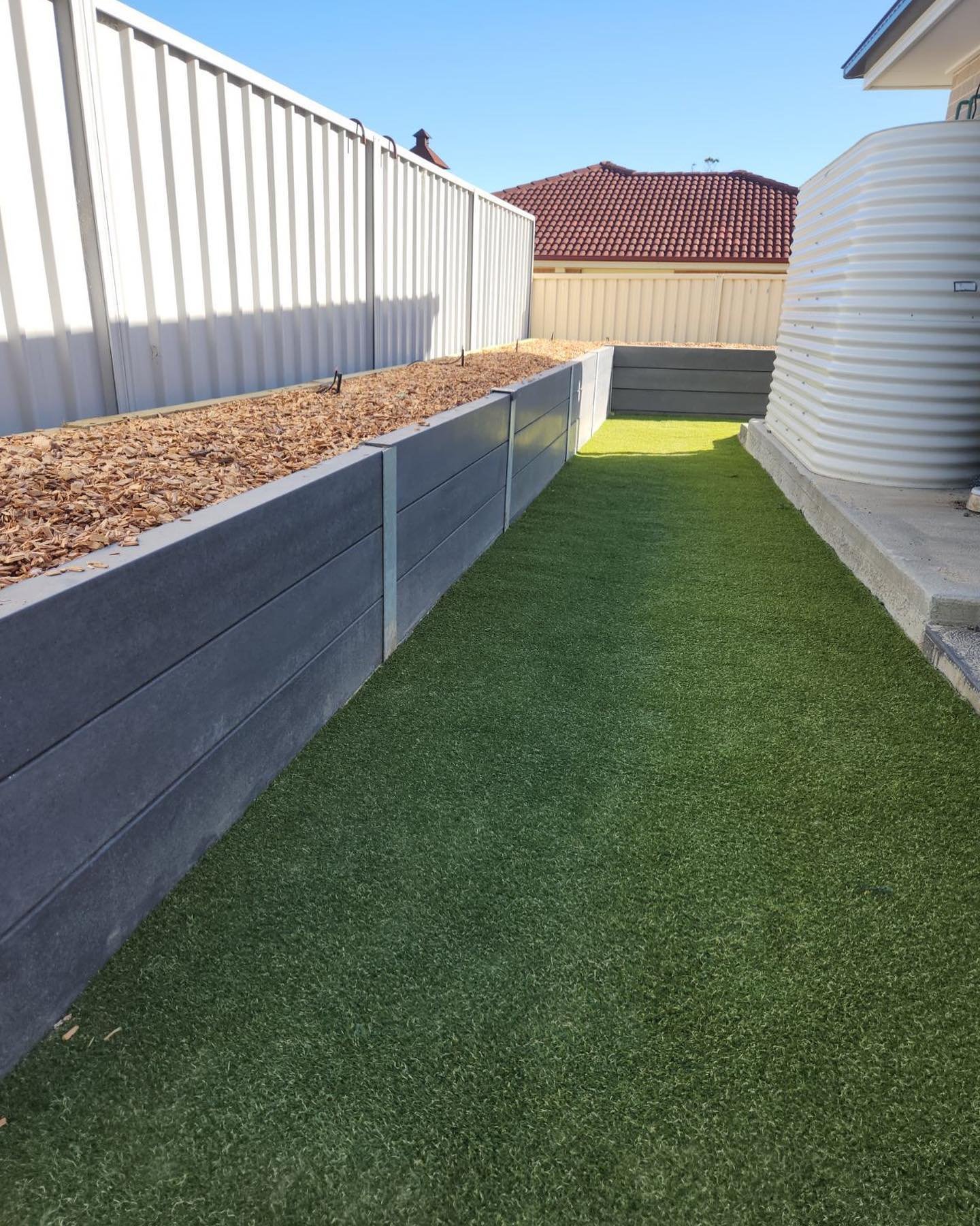 A backyard with artificial grass, a gray retaining wall filled with mulch, a white corrugated metal fence, and a white water tank. The sky is clear and blue.