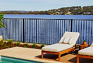 Poolside lounge chair and potted plants on a balcony overlooking a body of water with black pool fence.