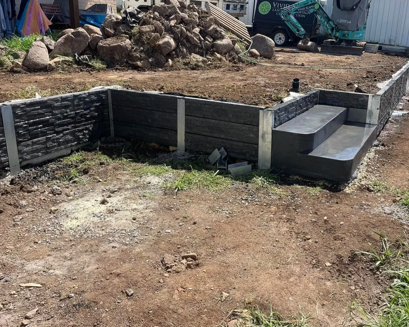 Construction site with a newly built retaining wall or garden bed with a small built-in step and concrete surface, surrounded by dirt and some grass, with construction equipment and vehicles in the background.