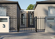 A black metal gate with vertical bars, flanked by two concrete pillars, in front of residential buildings.