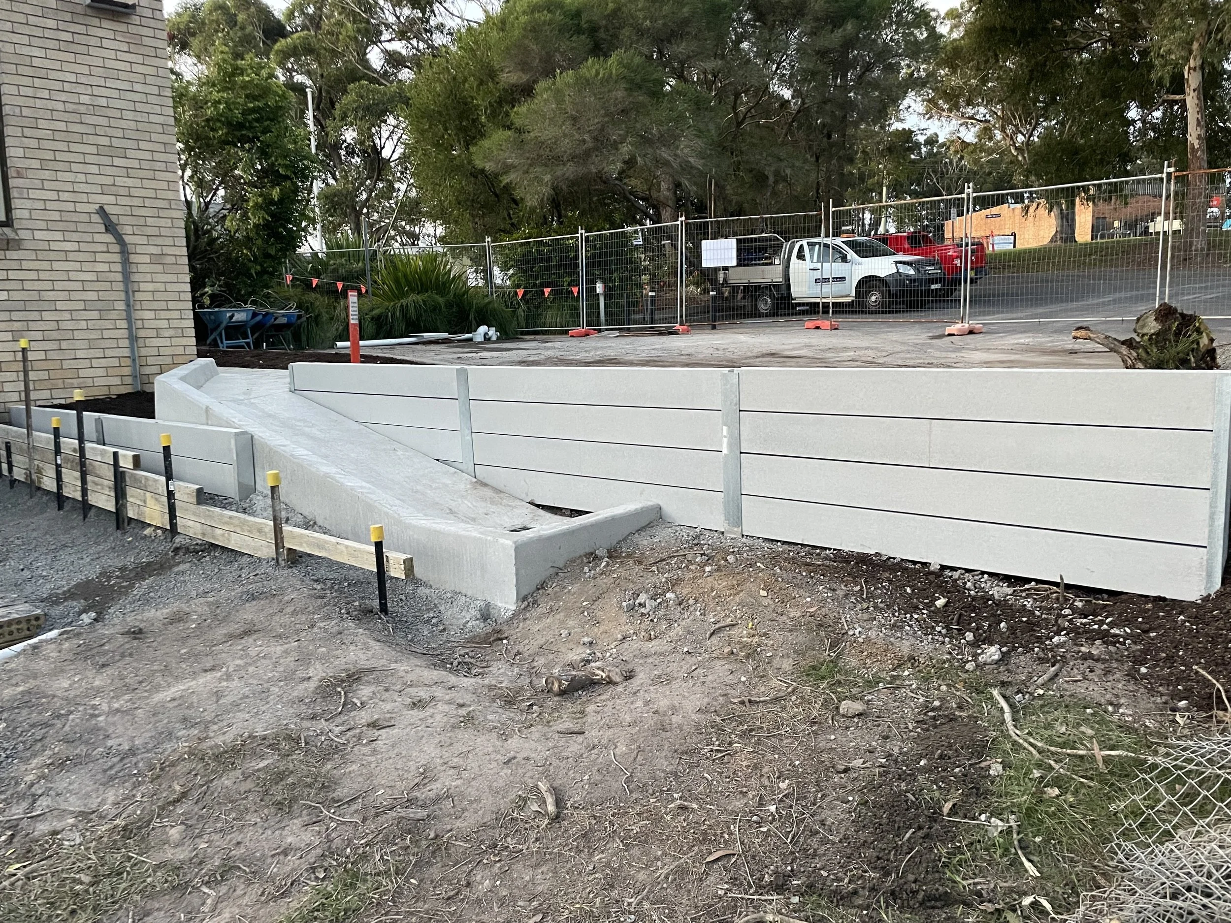 Newly constructed concrete retaining wall with a slope, installed next to a brick building and adjacent to a construction site with partial fencing and parked vehicles in the background.
