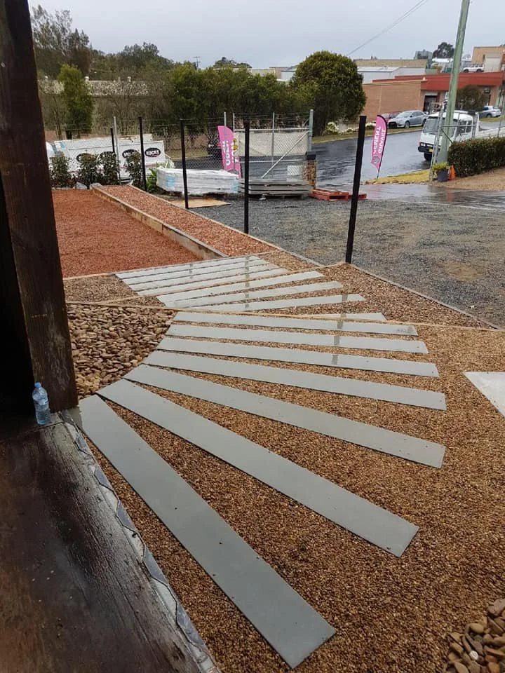 Newly installed curved concrete pavers on a gravel pathway outside, wet from rain, with construction materials and trucks in the background.