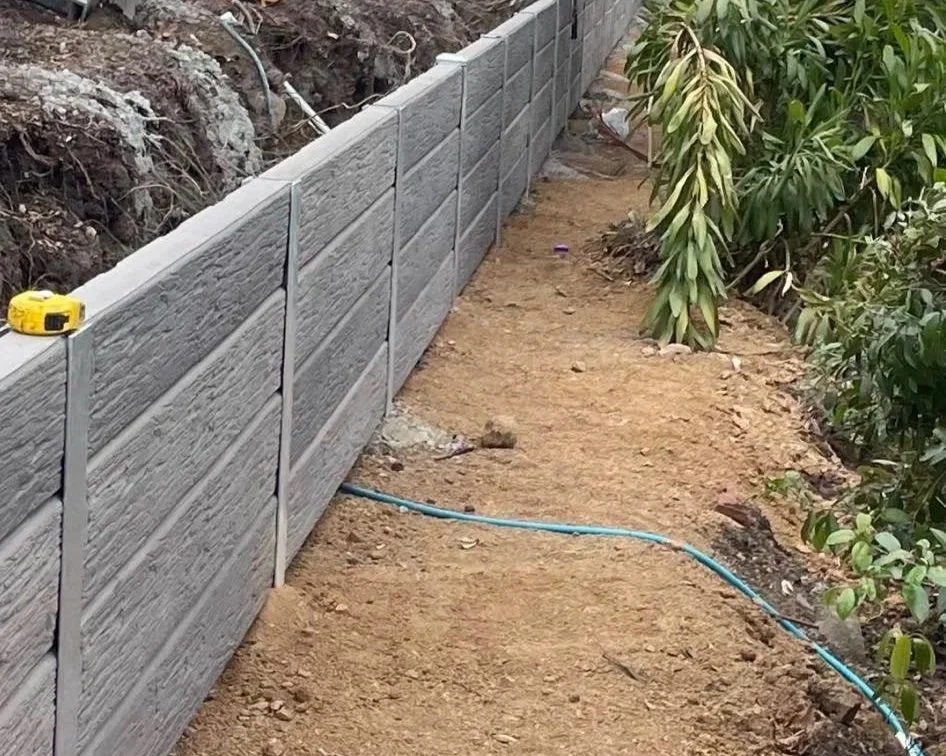 A newly built gray concrete fence on one side of a dirt pathway with tools and plants along the other side.