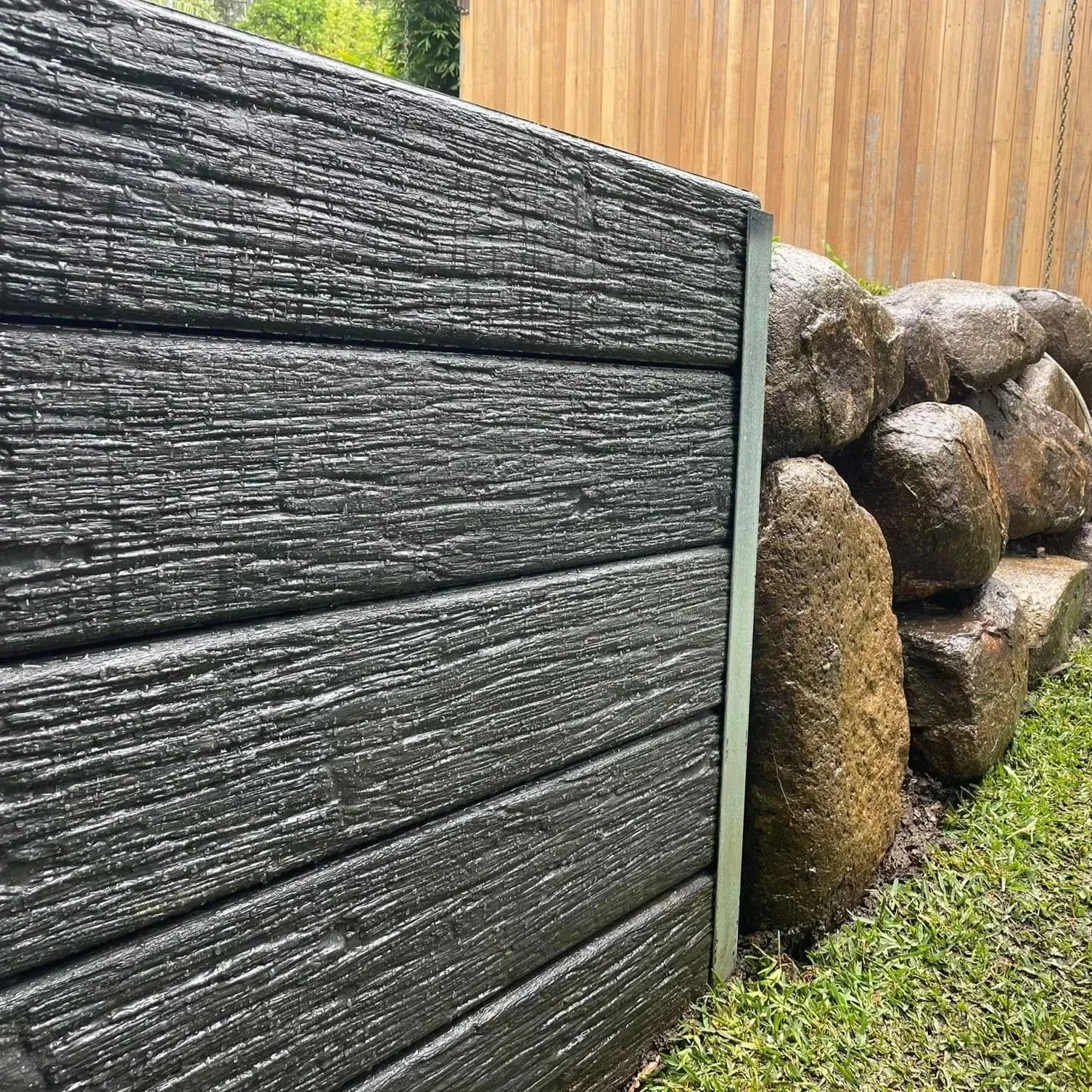 Close-up of black wooden fence panels with visible wood grain, alongside large brown rocks and green grass.