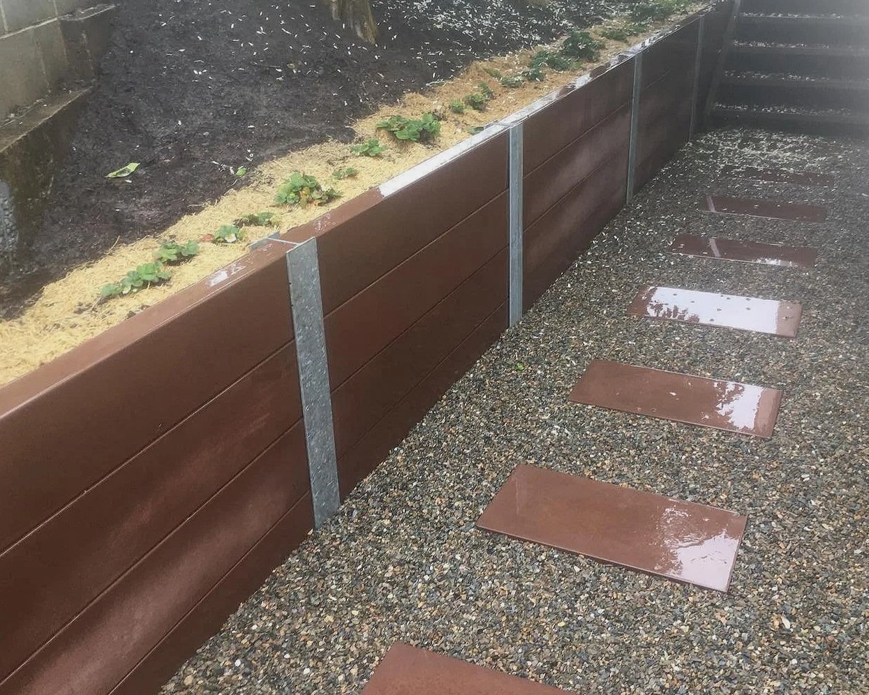 A rain-soaked outdoor path with rectangular brown stepping stones and a long brown wooden planter box with small plants and soil inside.