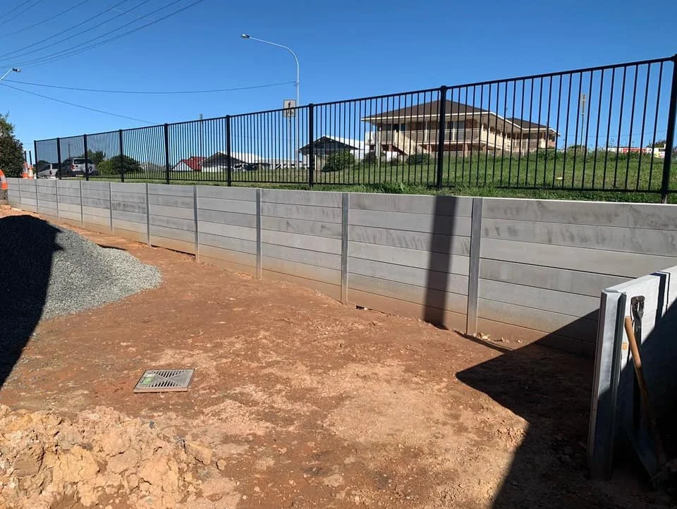 A construction site with a concrete retaining wall and black metal fence on top. The ground is dirt with a gravel pile on the left. In the background, there are houses and a blue sky.