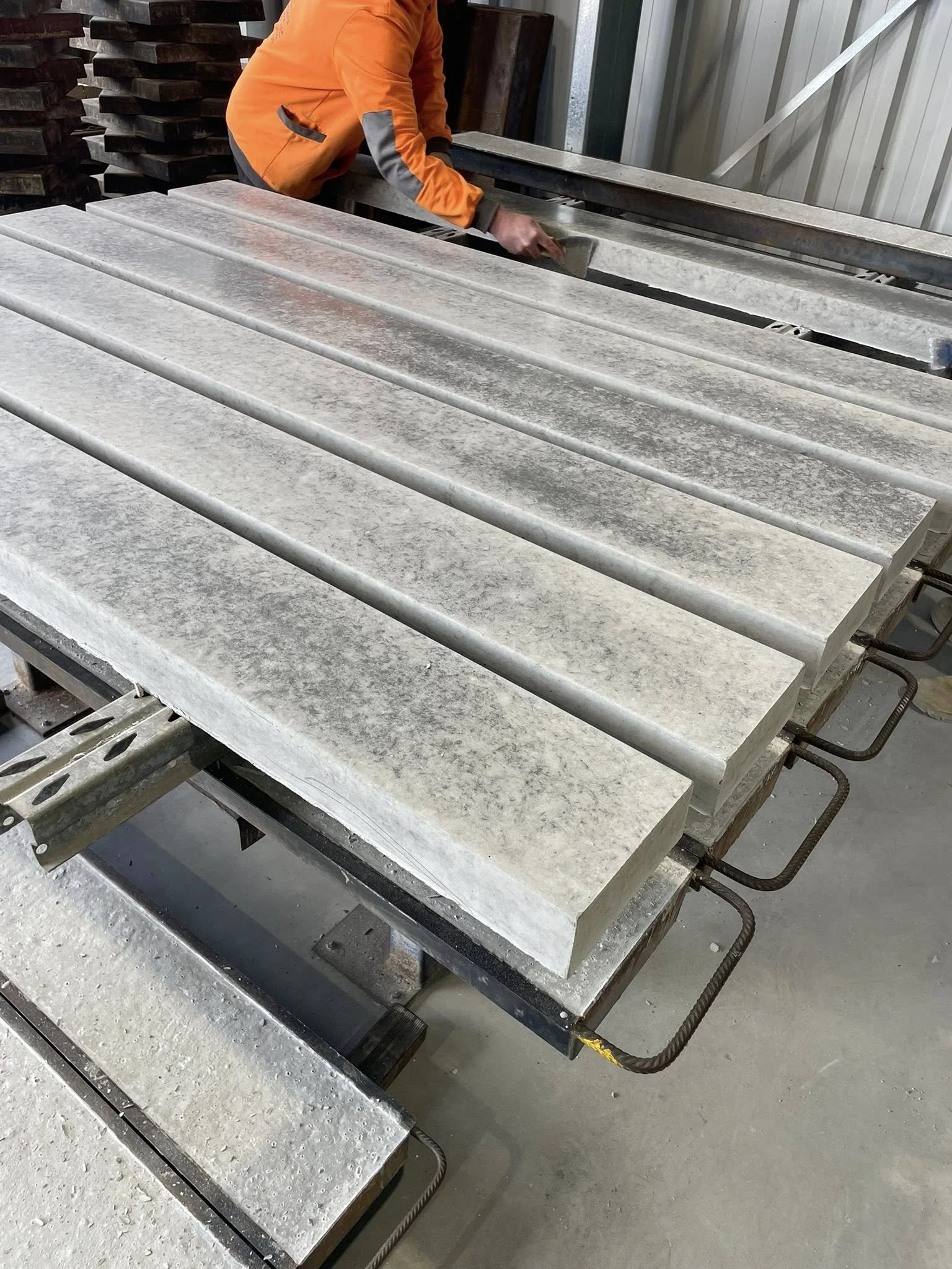 A worker in an orange jacket is smoothing the surface of a large, rectangular concrete sleeper with a trowel at a workshop.