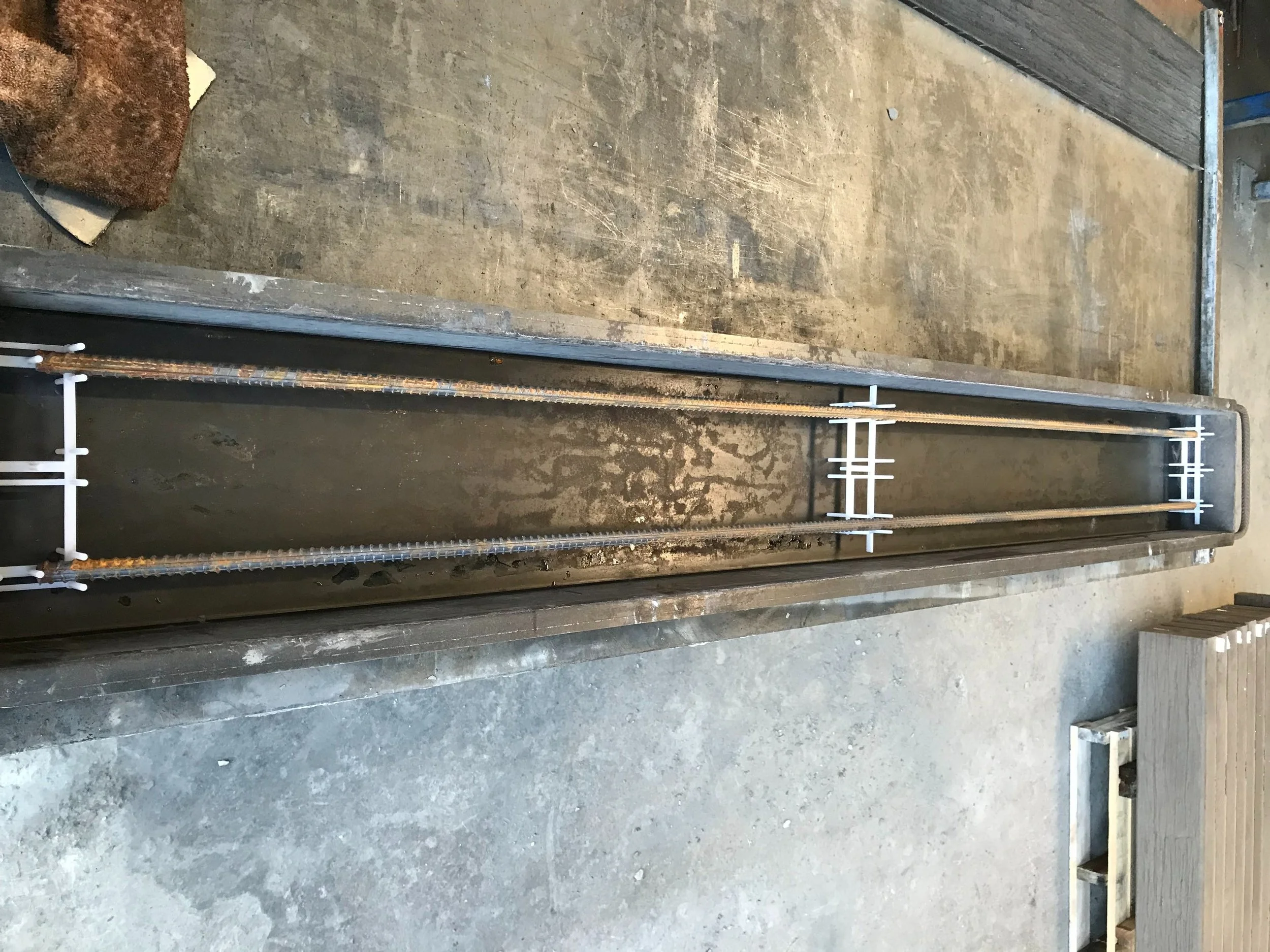 Long metal sleeper mould with two parallel metal rods inside a rectangular frame, supported by white plastic brackets, on a concrete floor in an industrial setting.