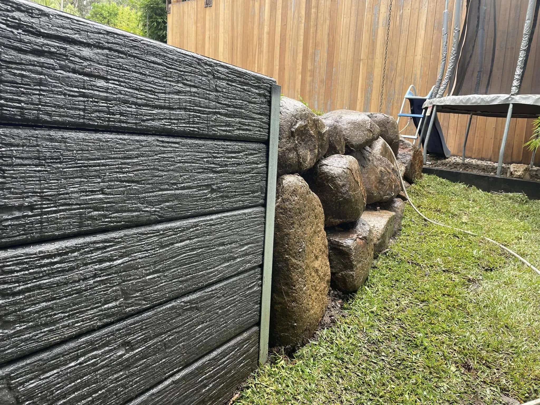 View of a backyard with a wooden fence, a stone retaining wall, and a grassy area with a trampoline and a slide in the background.