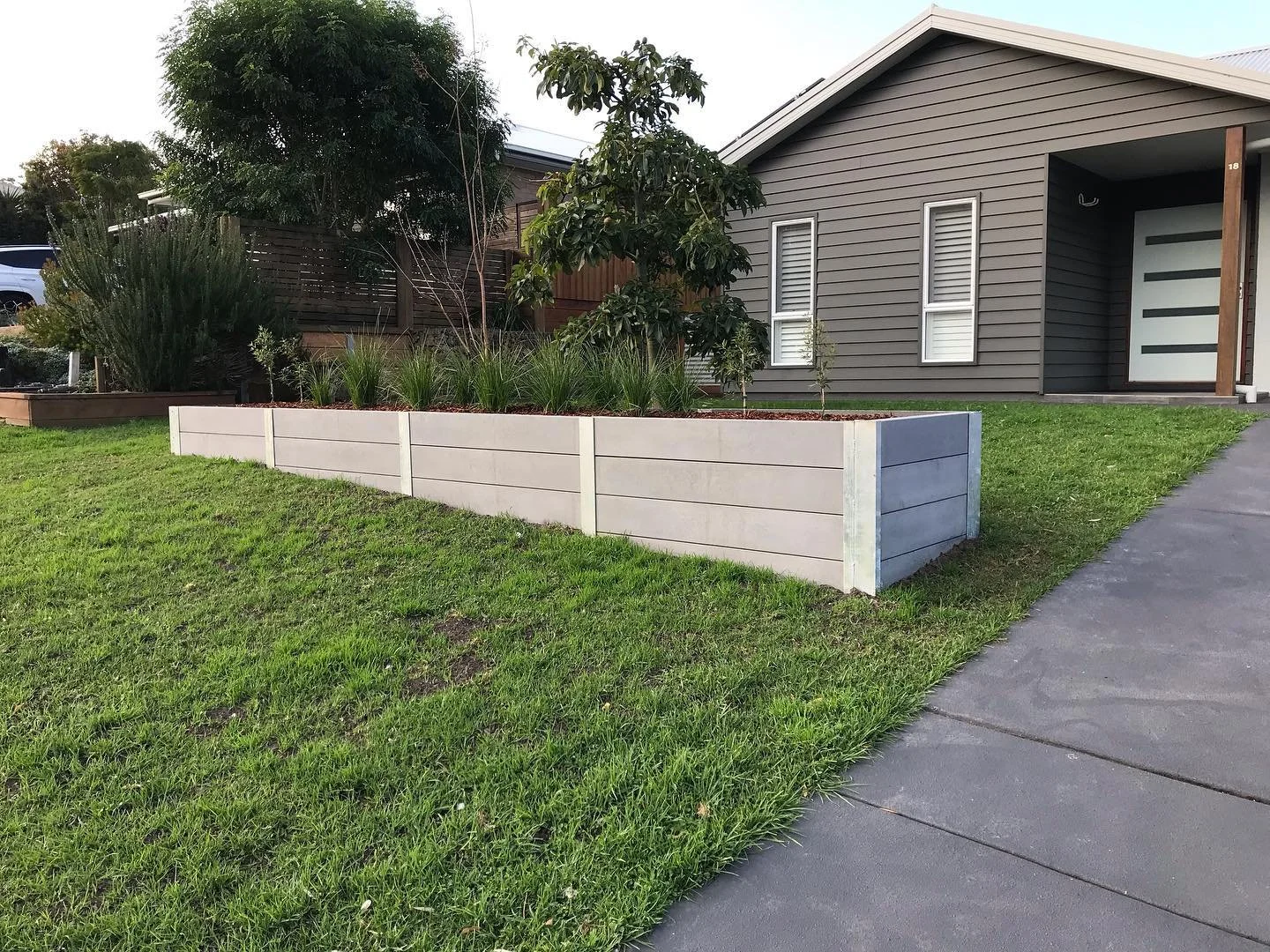 A modern house with gray siding, a white garage door with horizontal windows, and a small front yard. There is a concrete sidewalk and a raised white planter with green plants and trees in the yard.