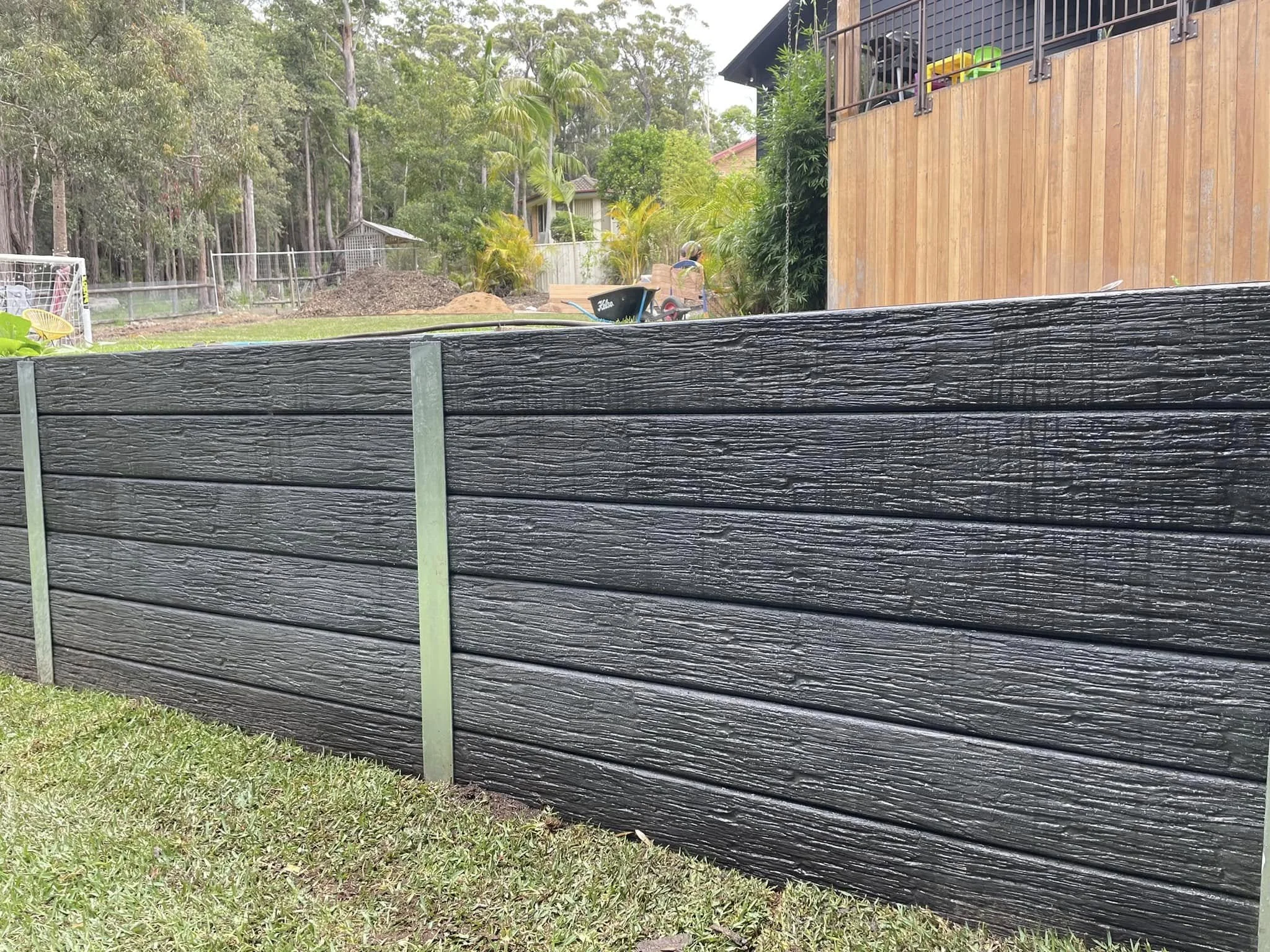 A black wooden fence in the foreground with a backyard that includes a play area, trees, and a wooden house or deck in the background.