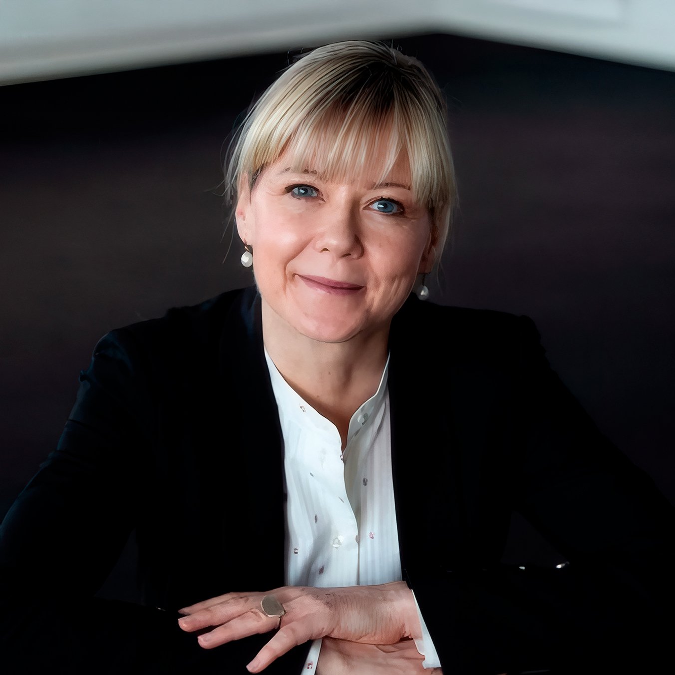 Et portræt af en kvinde med blond hår og blå øjne, iført en hvid skjorte og sort blazer, smilende og sidder ved et bord.