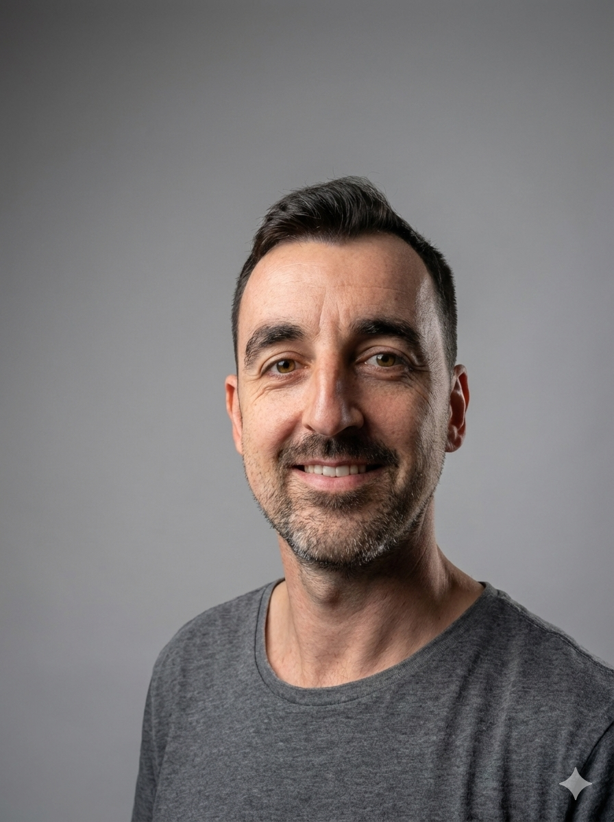 Portret van een man met donkere korte haar en een stoppelbaard, lachend, tegen een effen grijze achtergrond, gekleed in een grijs T-shirt.