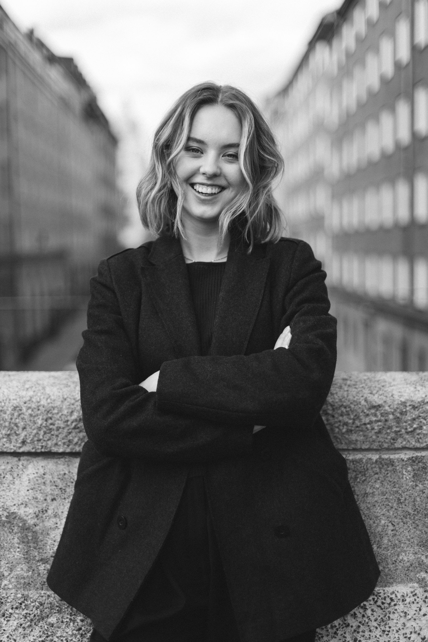A smiling woman with wavy blonde hair standing outdoors with her arms crossed, wearing a dark blazer and a black top, in front of a cityscape with multi-story buildings.