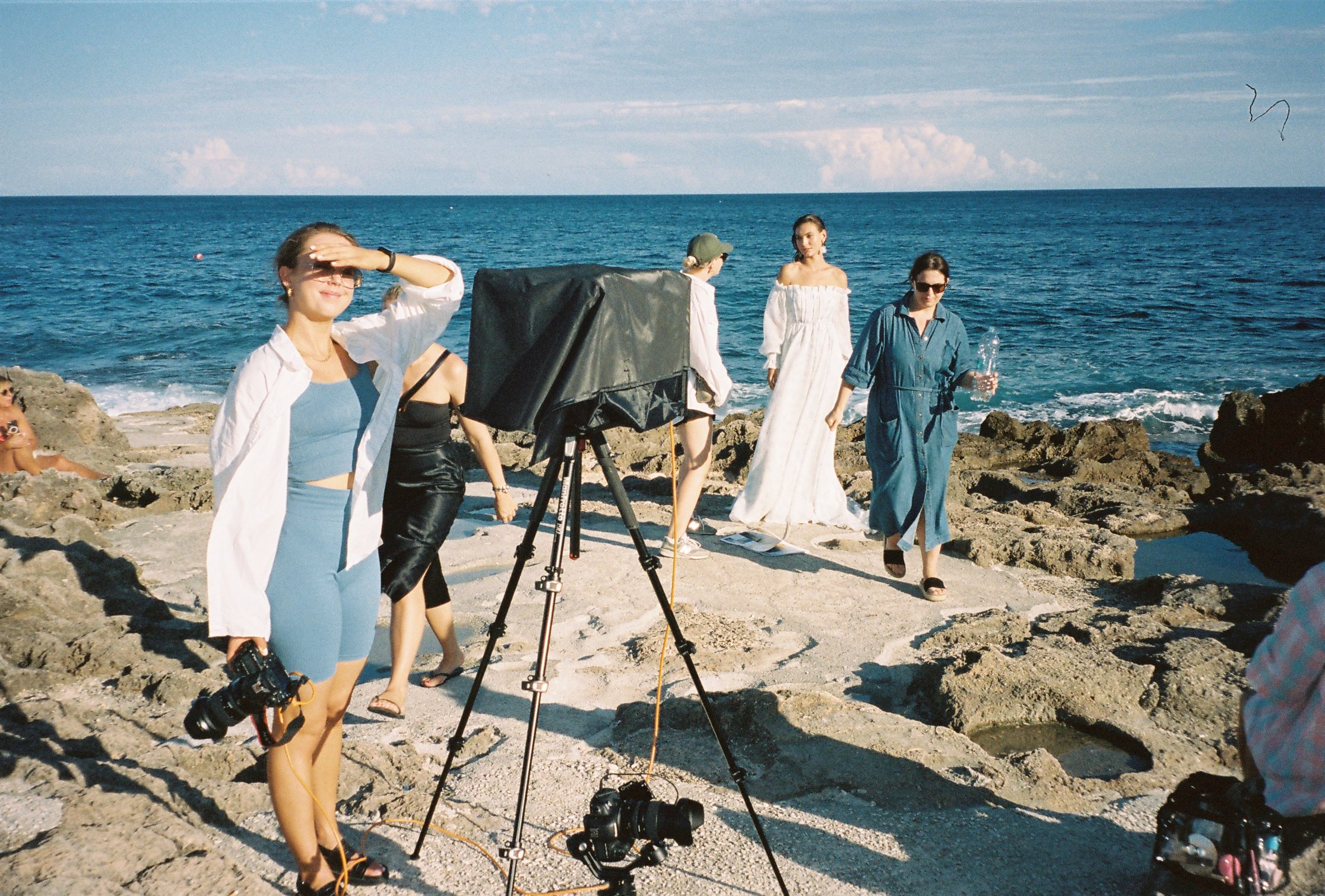 Group of women on rocky beach during a photoshoot with a camera and lighting equipment, ocean in background.