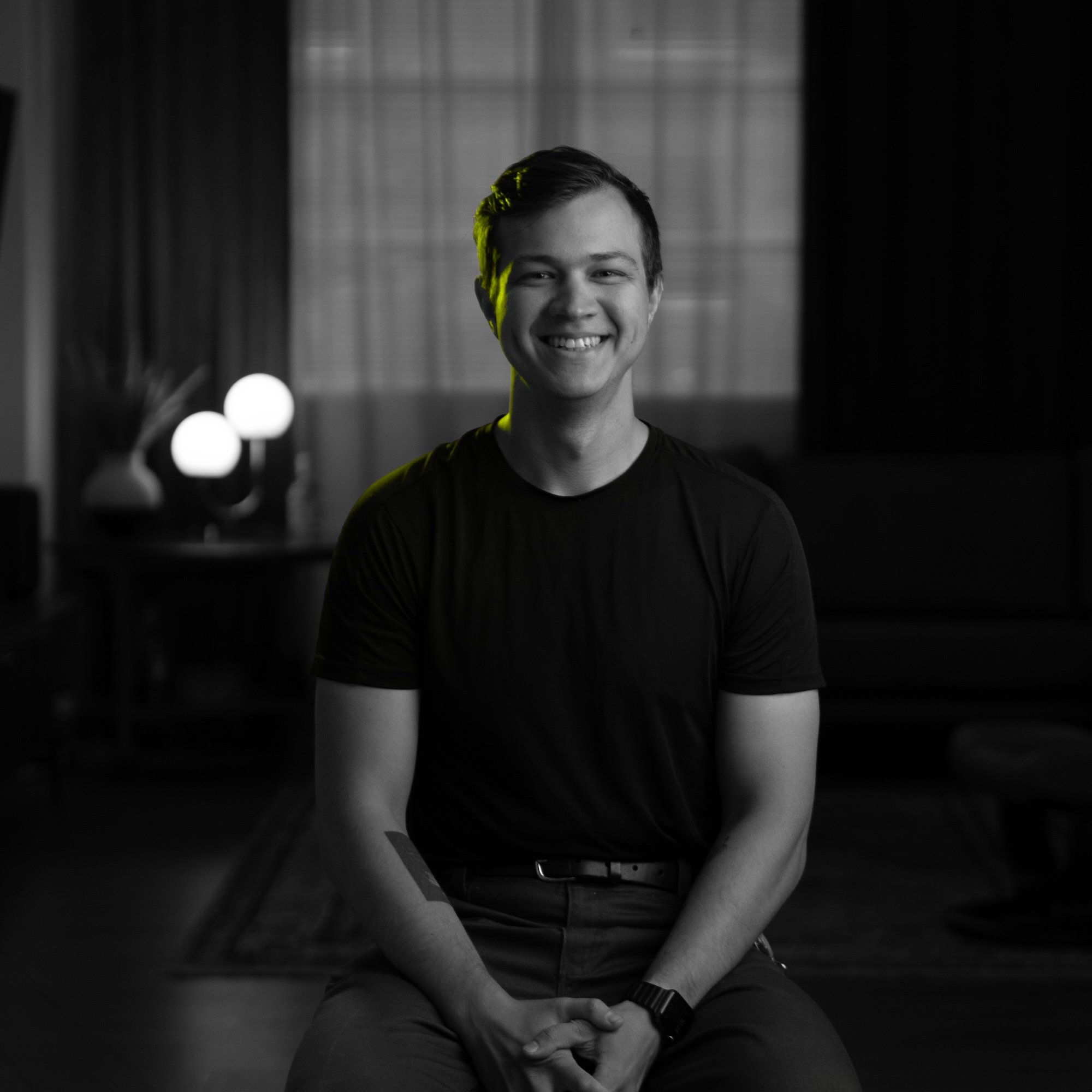 A young man smiling while sitting indoors in a dimly lit room with modern decor and soft lighting.