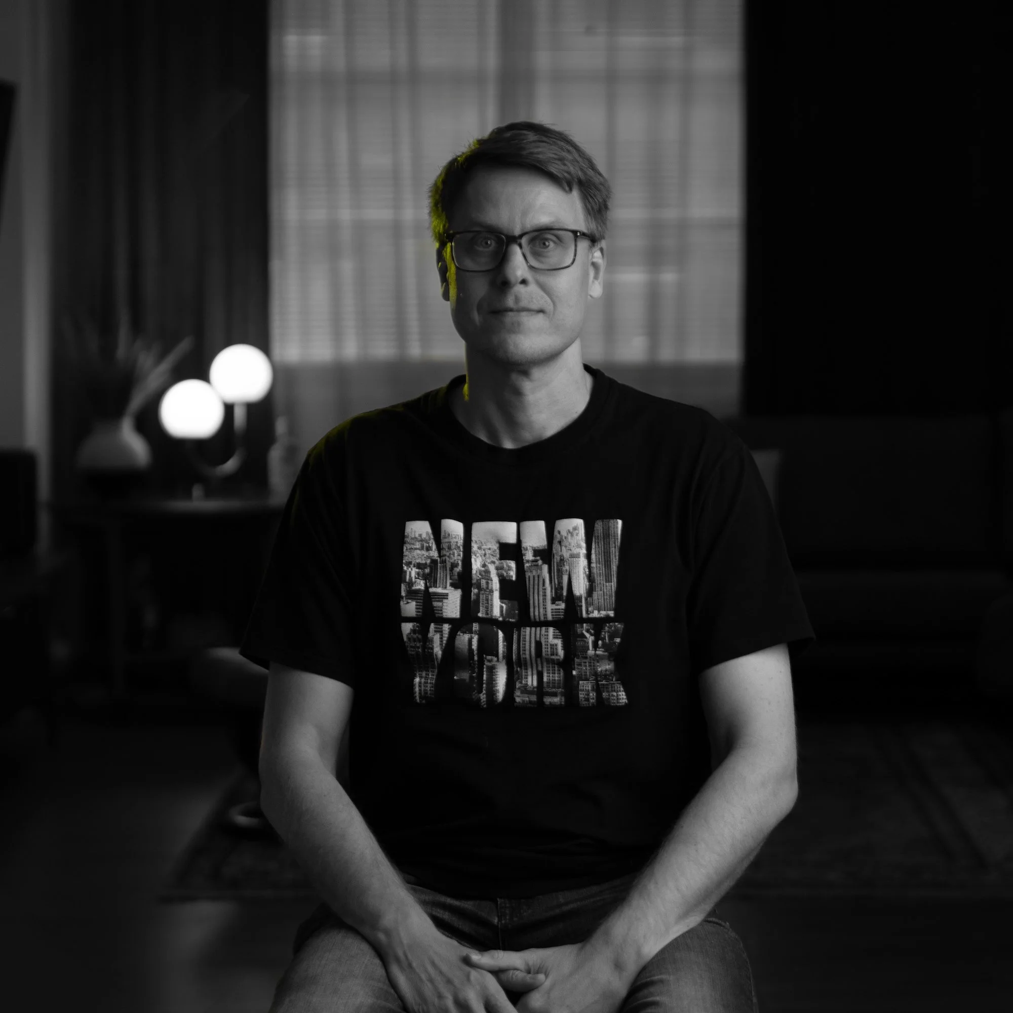 A man with glasses and short hair sitting on a chair in a dimly lit room with modern decor, including a couch and lamps in the background.