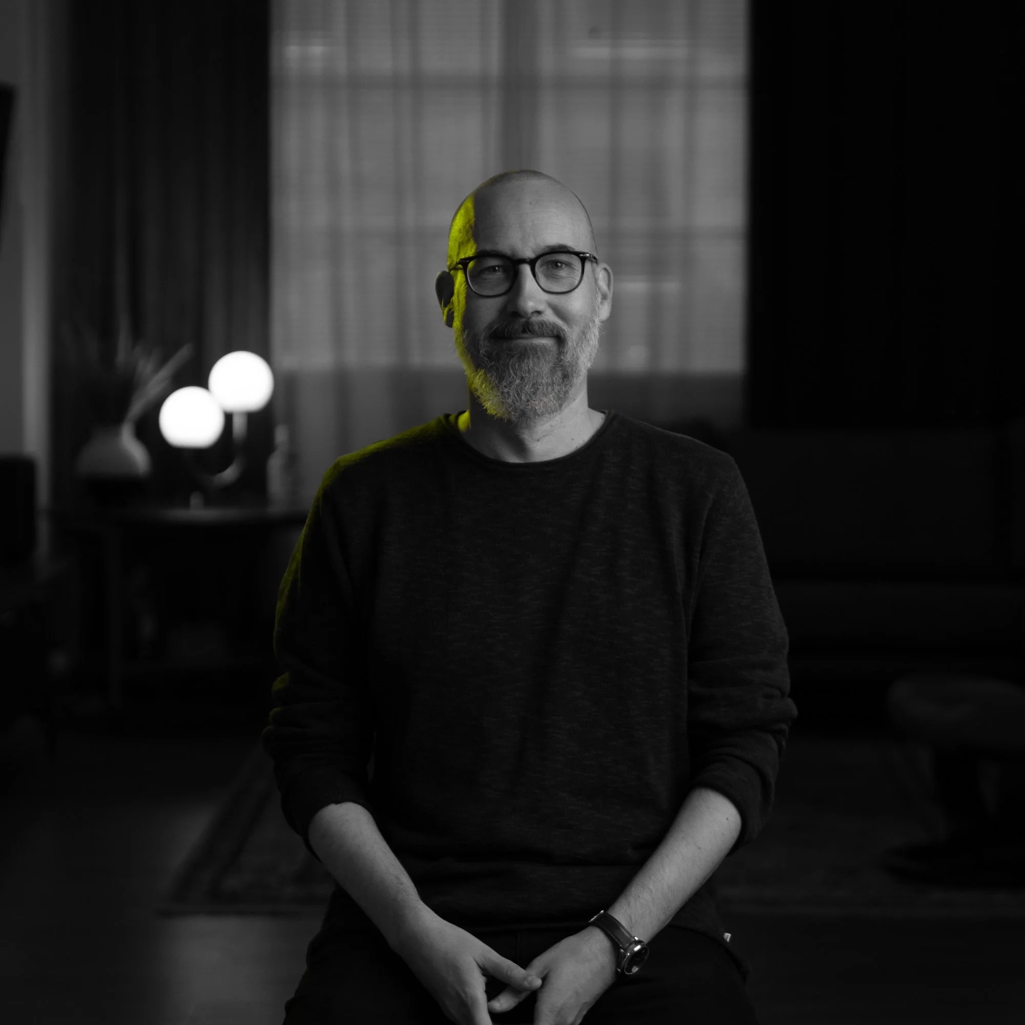A man with glasses and a beard sitting in a dimly lit room with curtains and lamps in the background.