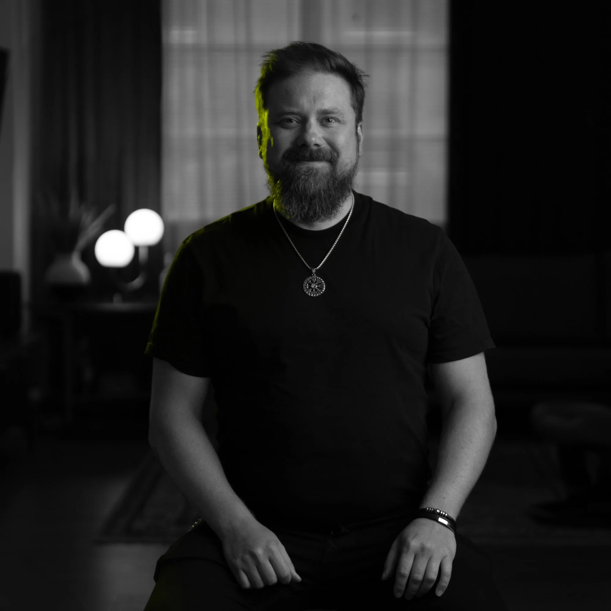 A smiling man with a beard and mustache wearing a black t-shirt, a necklace with a circular pendant, and a bracelet, sitting indoors with a blurred background, black and white with a subtle green accent on the left side of his hair.