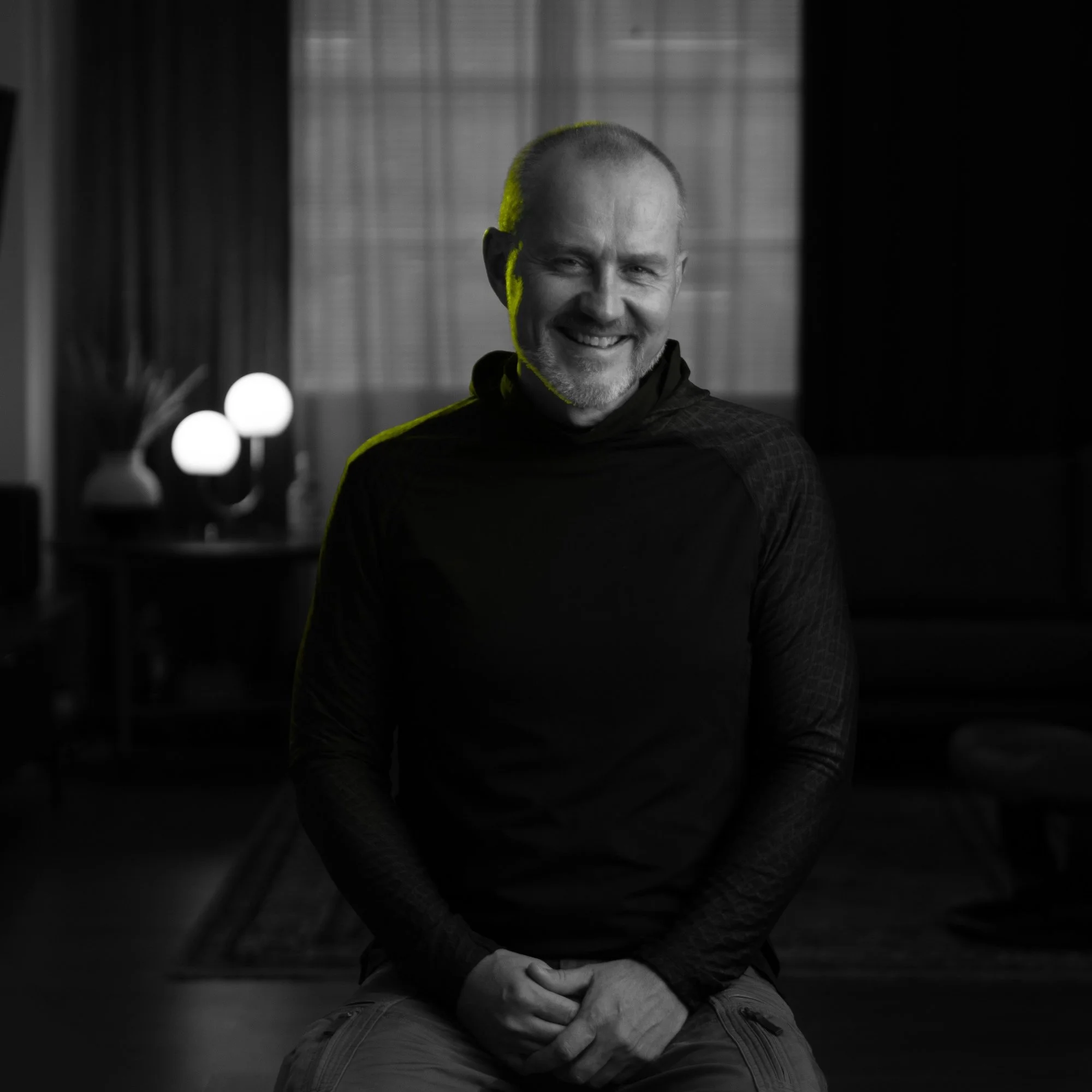 Black and white photo of a middle-aged man sitting indoors, smiling, with a lamp and curtains in the background.