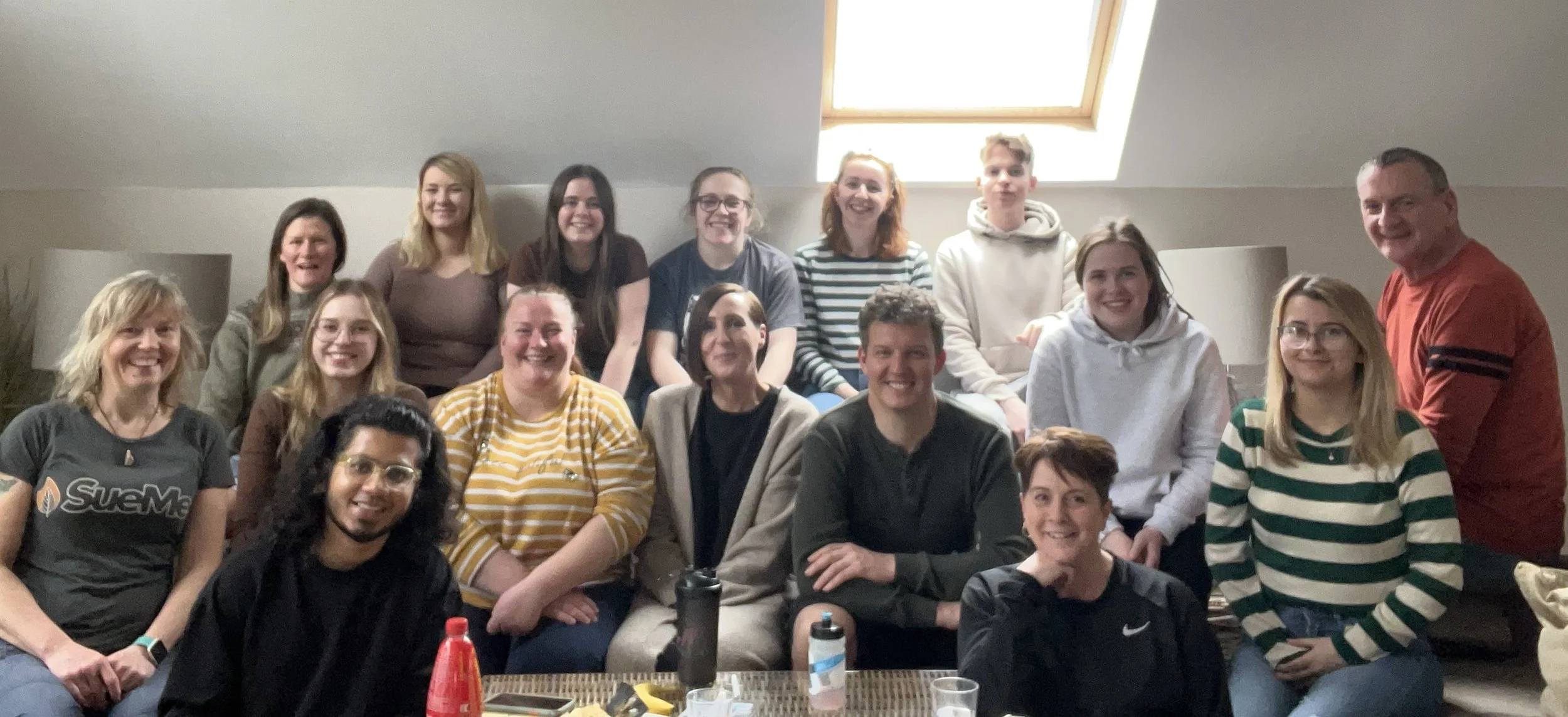 A group of people smiling and posing together indoors, some seated and others standing, with bright natural light coming through a skylight.