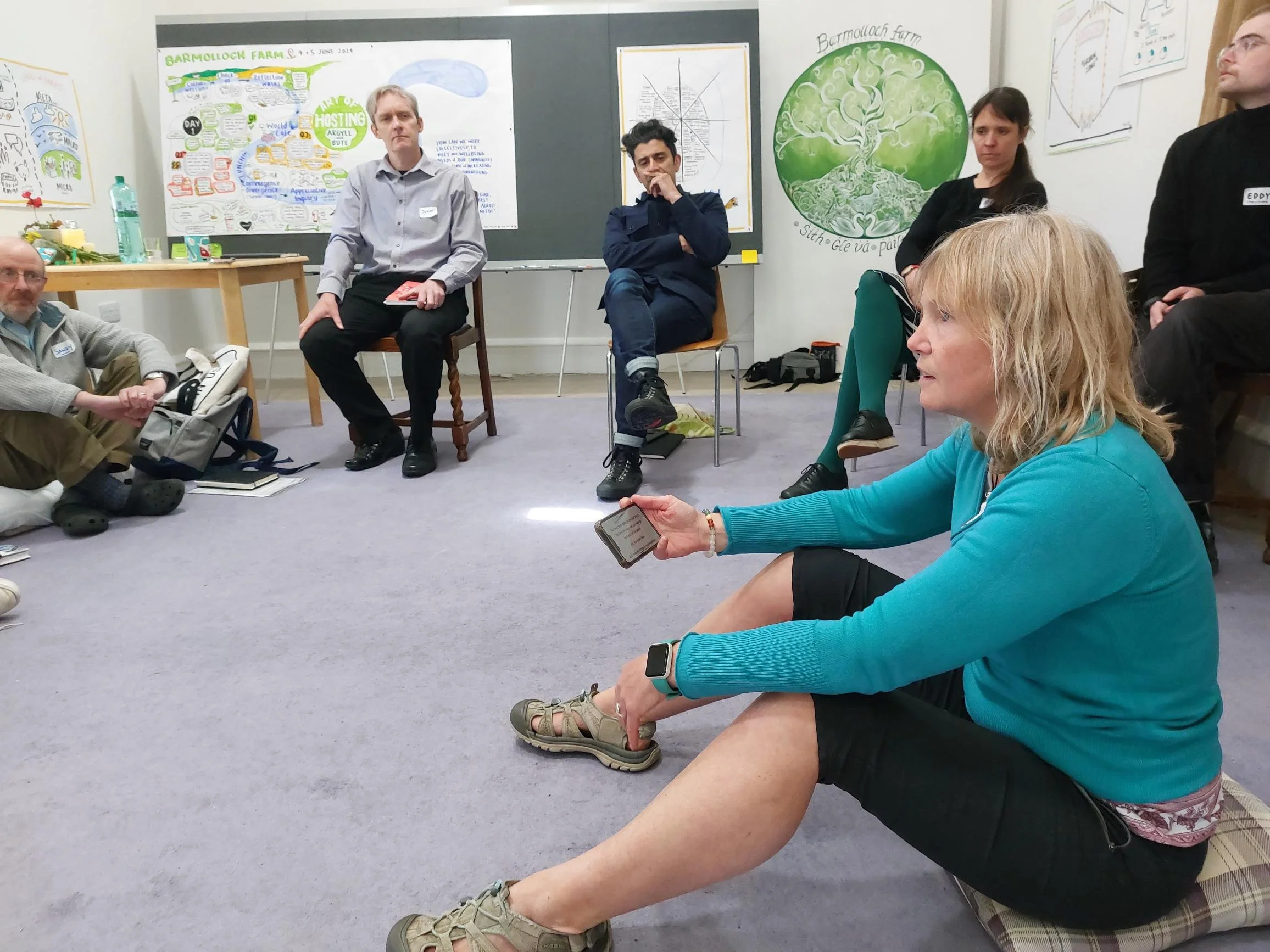 Group of people sitting in a circle on the floor, engaged in a discussion. The room has posters with diagrams on the walls. A woman in a turquoise sweater holds a phone, speaking.