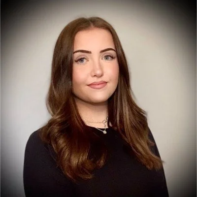 A young woman with long, wavy brown hair, wearing a black top and a necklace, posing against a plain background.