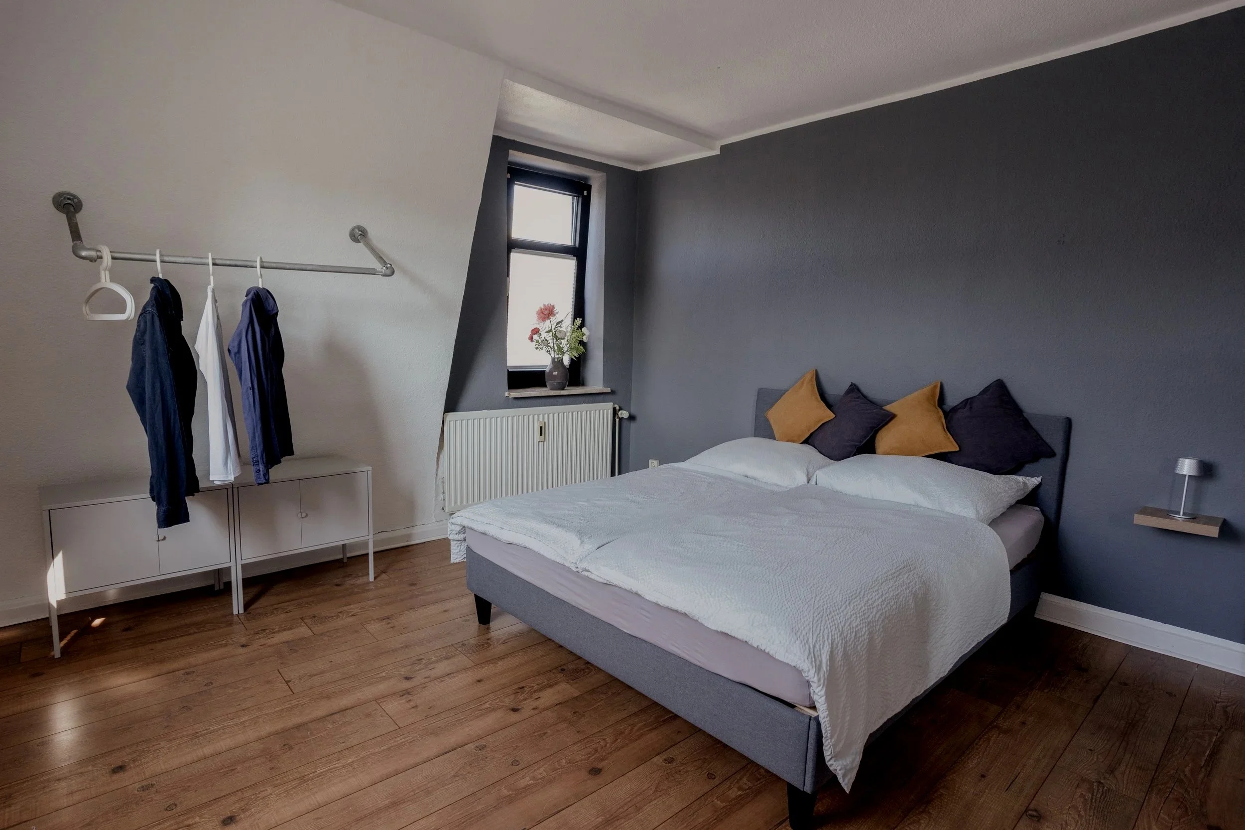 Ein Schlafzimmer mit grauer Wand, einem Doppelbett mit weißen Bettzeug und vier Kissen in Grau und Braun, einem Fenster mit Blumen, einem weißen Möbelstück mit Kleidung und einem kleinen Nachttisch mit Lampe.